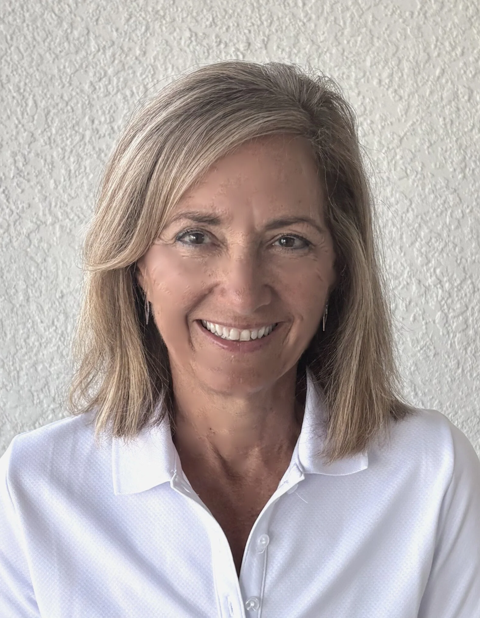 A woman with shoulder-length blonde hair smiling in front of a textured white wall, wearing a white collared shirt.
