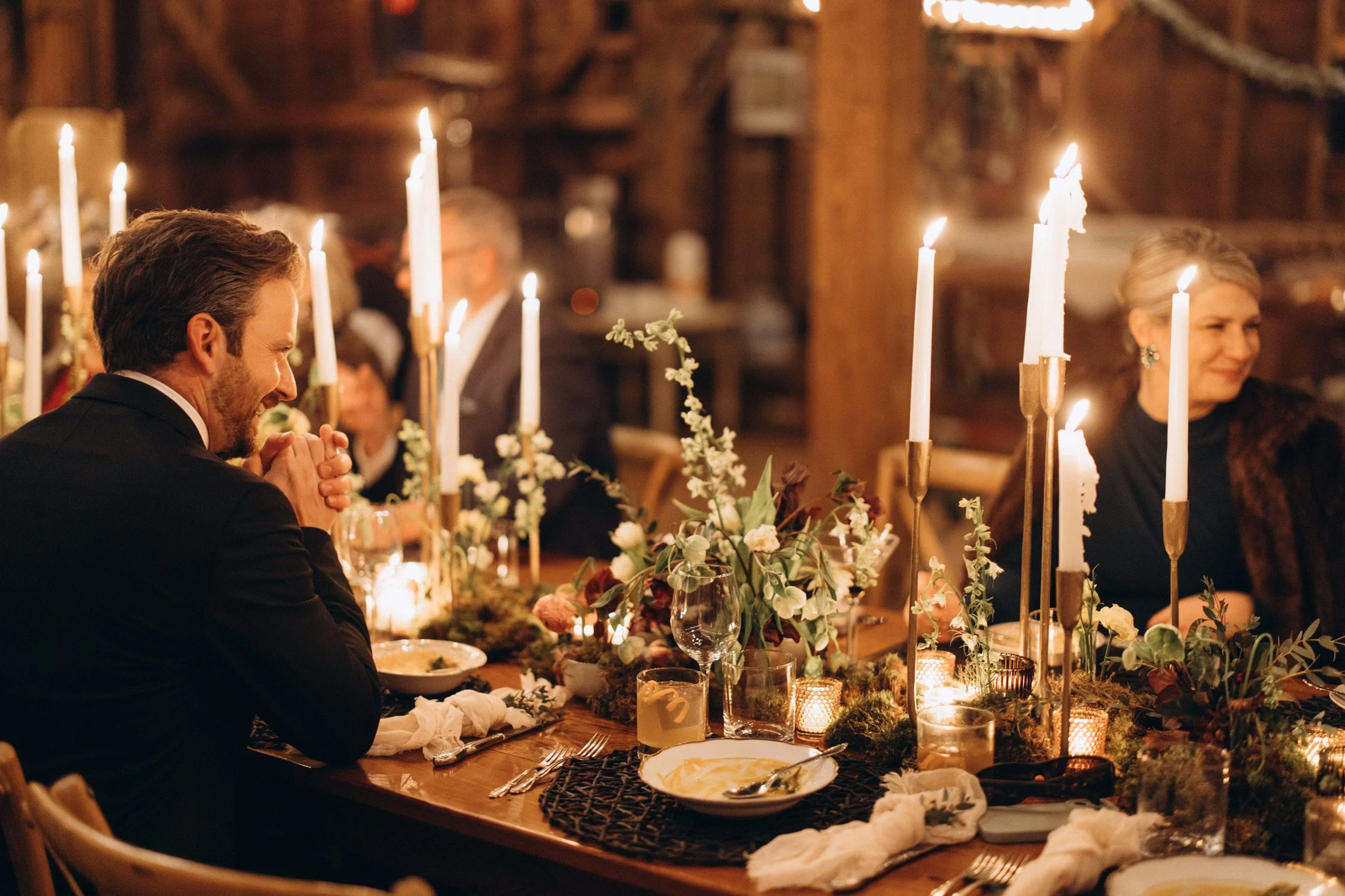 A man with dark hair and a beard, dressed in a tuxedo, is sitting at a candlelit dinner table with a woman with blonde hair and earrings, both smiling. The table is decorated with tall white candles, flowers, and candles in glass holders.