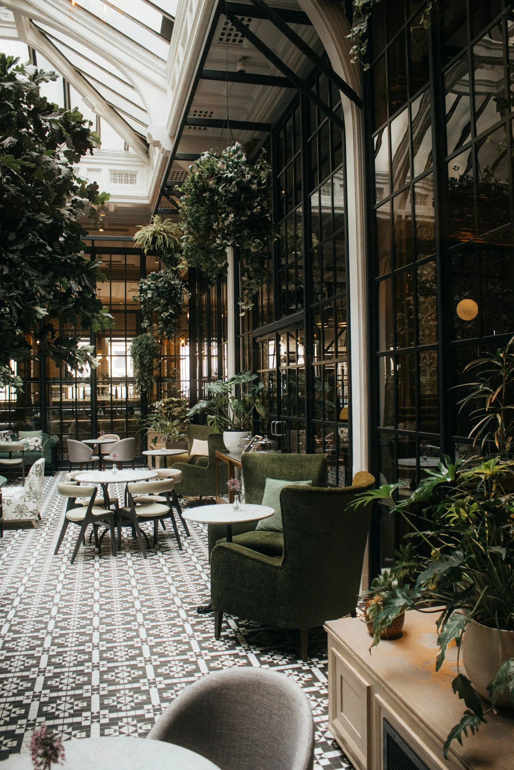 Interior of a stylish restaurant or cafe with black-framed glass walls, hanging plants, and a mix of modern and vintage furniture, including armchairs and small tables, with patterned tile flooring and large windows providing natural light.
