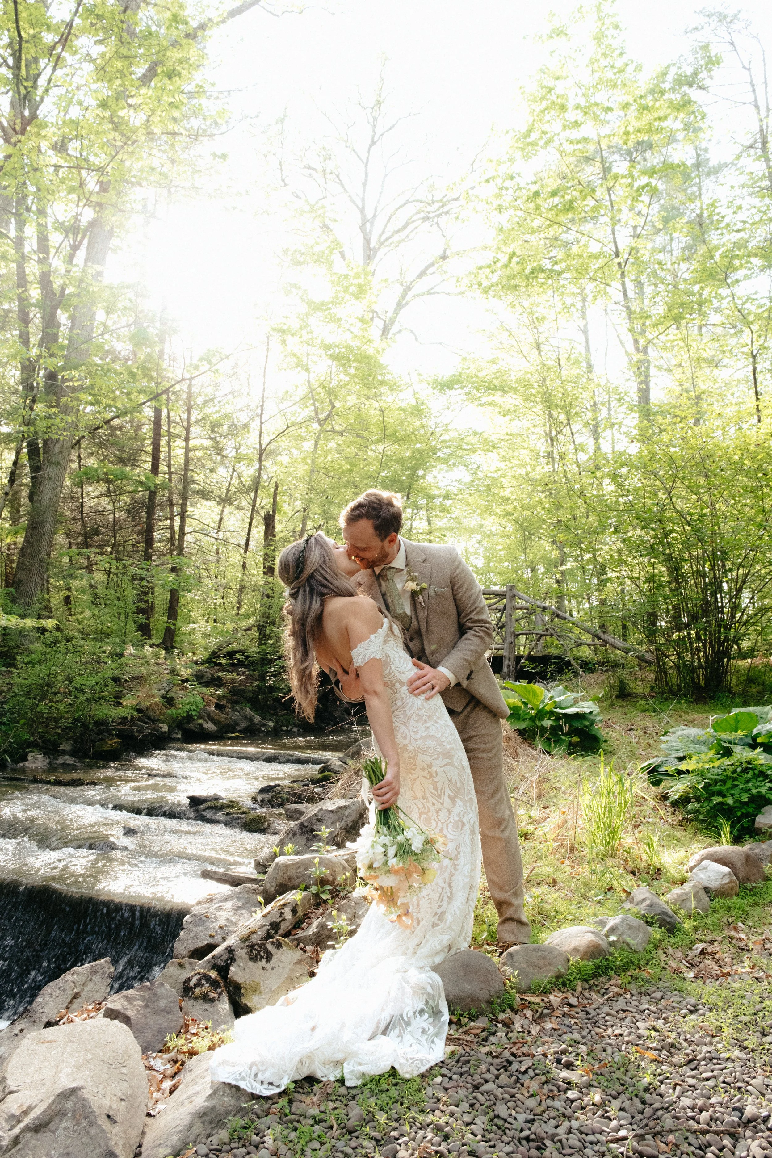 A couple in wedding attire standing by a creek in a lush green forest, sharing a kiss. The bride holds a bouquet of white flowers, and the groom is wearing a beige suit. Sunlight filters through the trees.