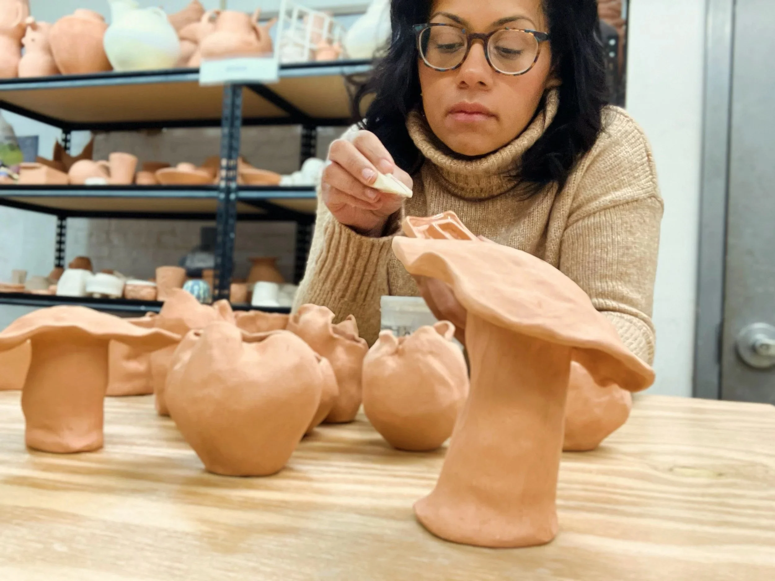 Woman wearing glasses examining ceramic figurines on a wooden table, with shelves of similar pottery in the background.
