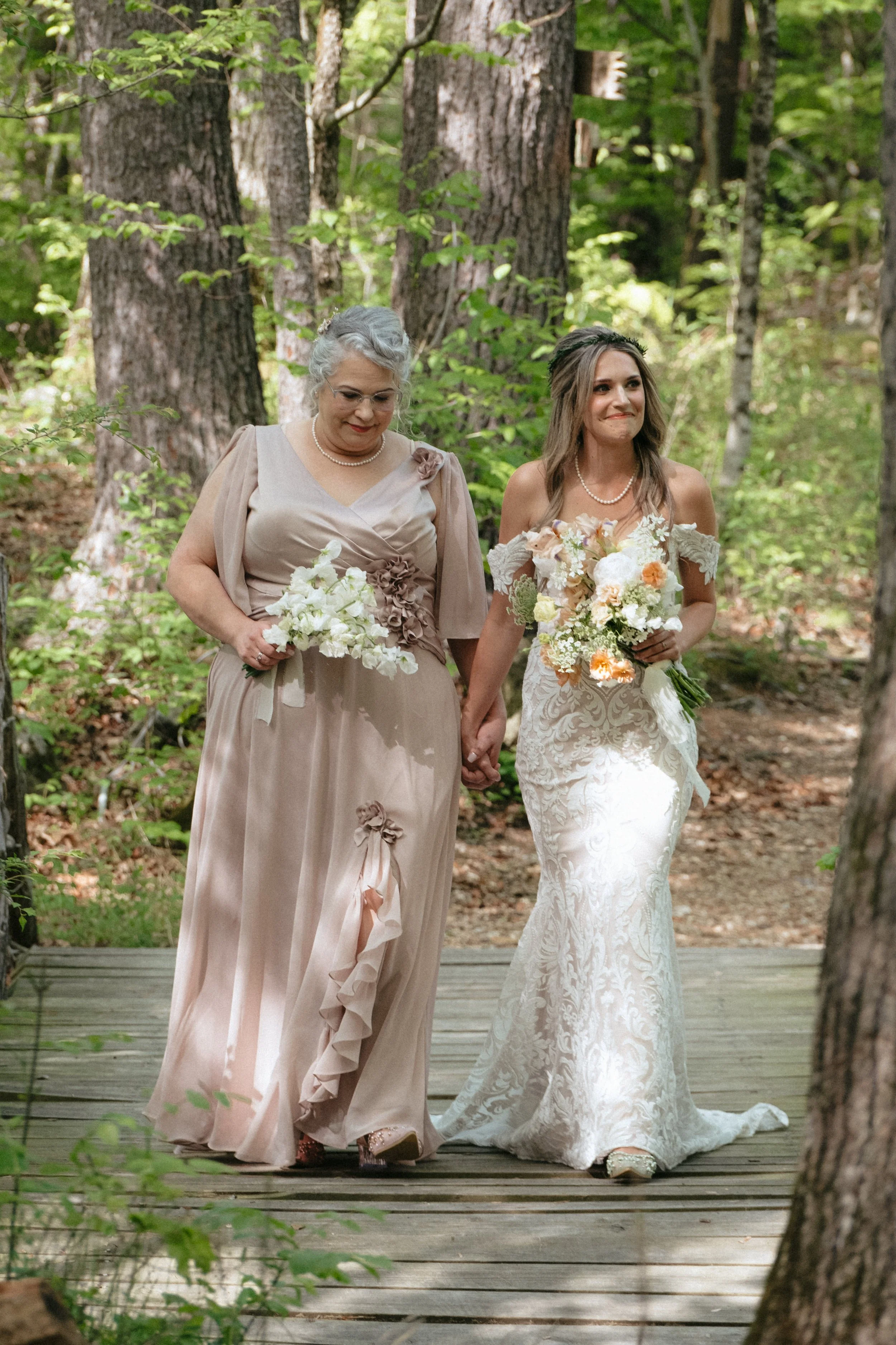 A bride and an older woman, possibly her mother, holding hands and walking through a wooded outdoor setting during a wedding ceremony. The bride wears a lace wedding gown and holds a bouquet of flowers; the older woman wears a champagne-colored dress and holds a bouquet.