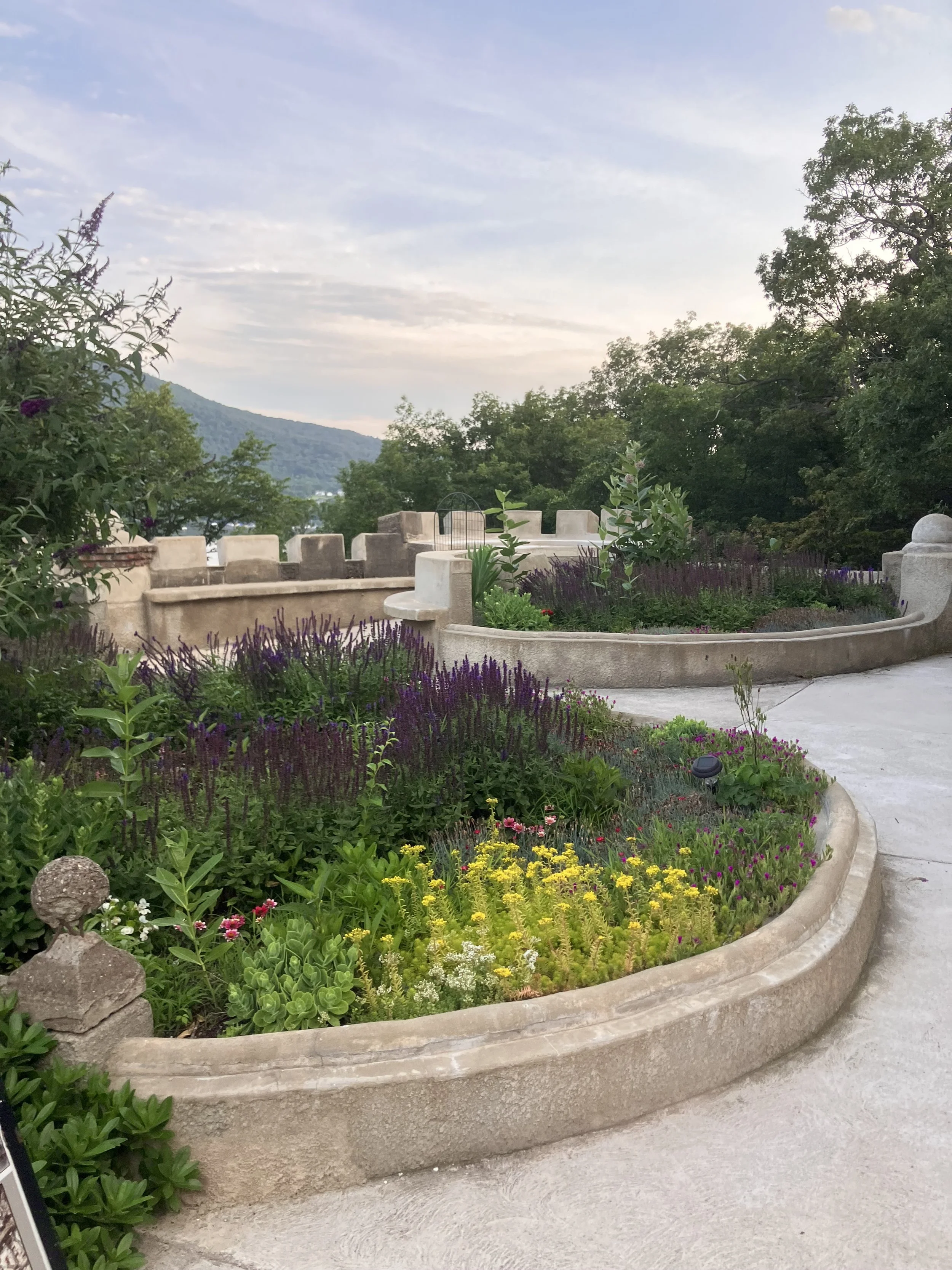 A circular garden bed at Bannerman Island filled with colorful flowers and green plants, surrounded by a concrete border, with trees and a mountain in the background under a partly cloudy sky.