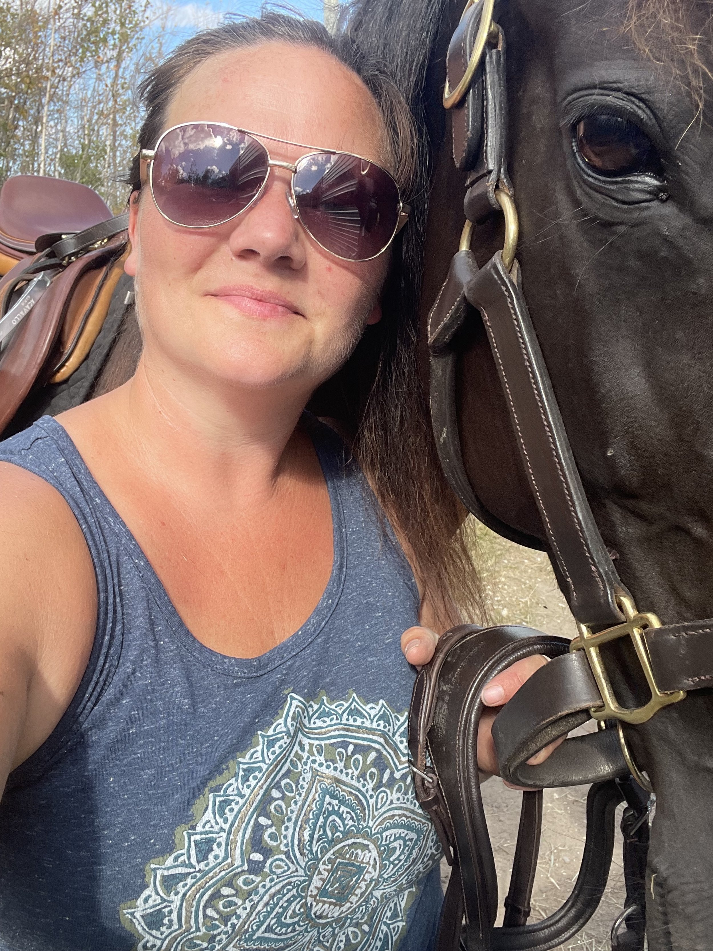 A woman with sunglasses, wearing a blue tank top, taking a selfie with a black horse, which is bridled and saddled.