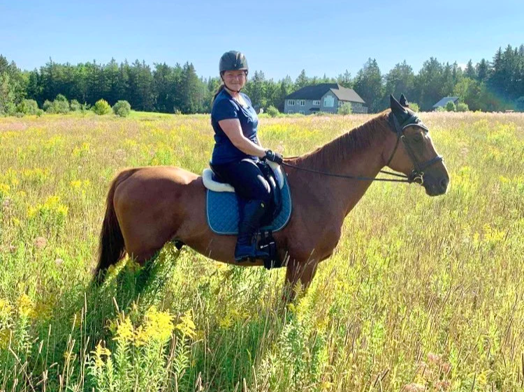 A woman riding a brown horse in a grassy field with yellow flowers, green trees, and a house in the background on a sunny day.