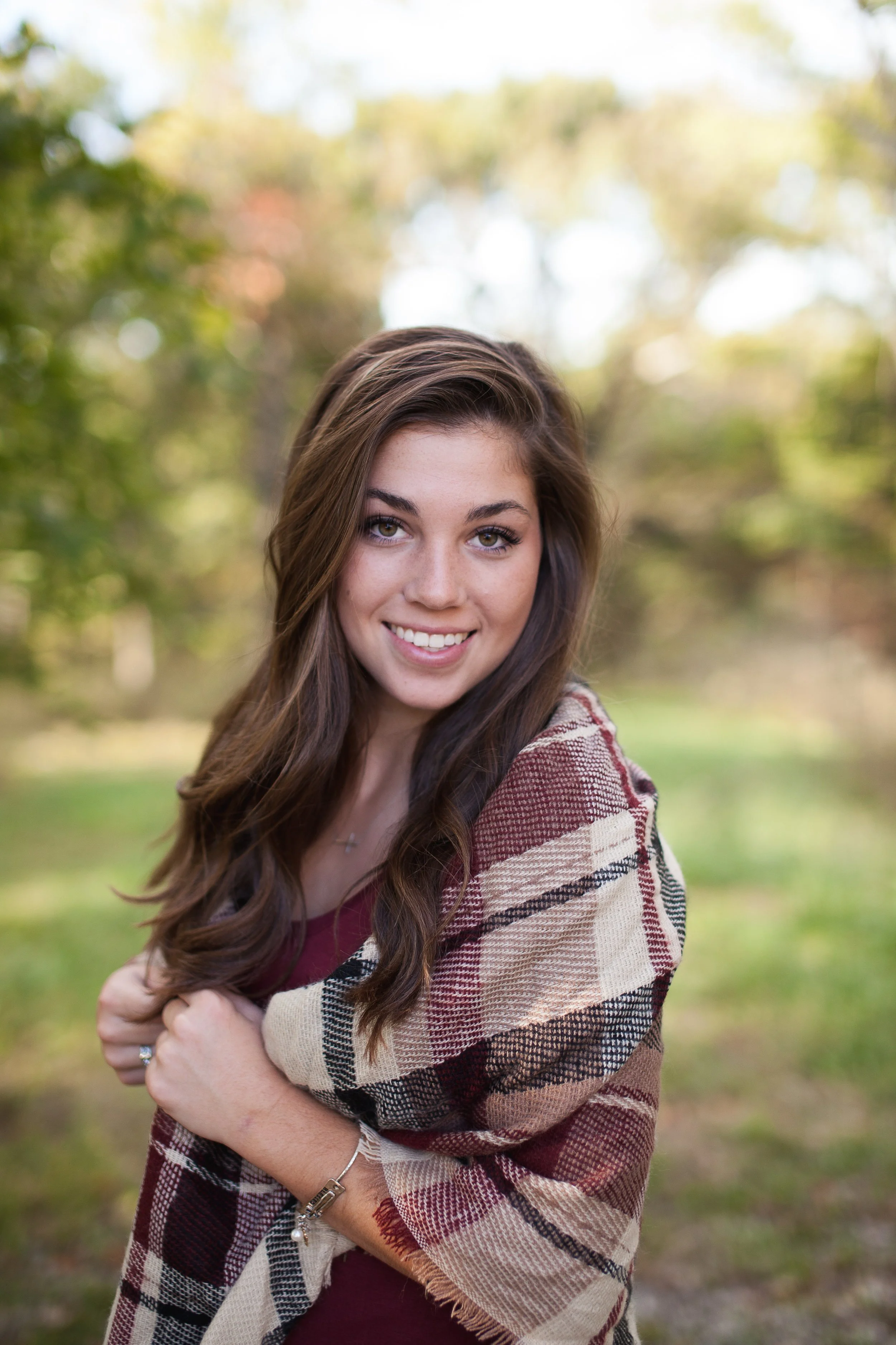 A young woman with long brown hair smiling outdoors, wrapped in a plaid shawl in a park with autumn-colored trees in the background.