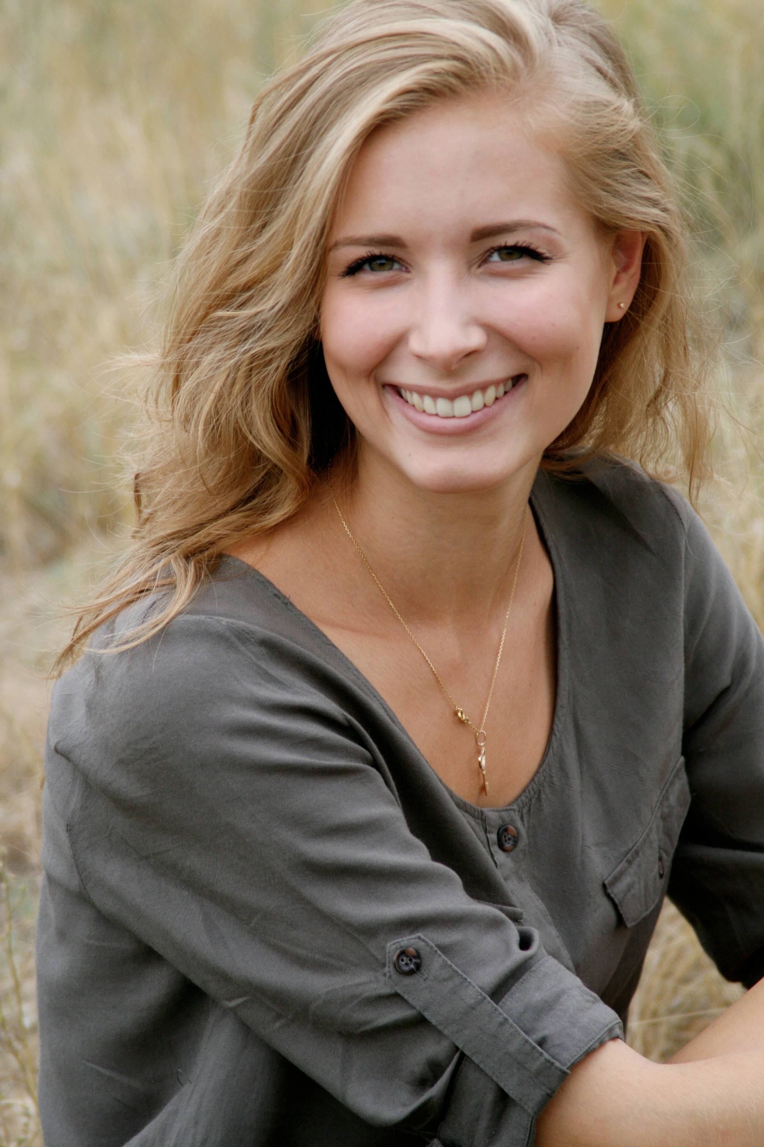 A young woman with blonde hair, smiling, wearing a dark gray shirt, and a gold necklace with a small pendant, outdoors in a field.