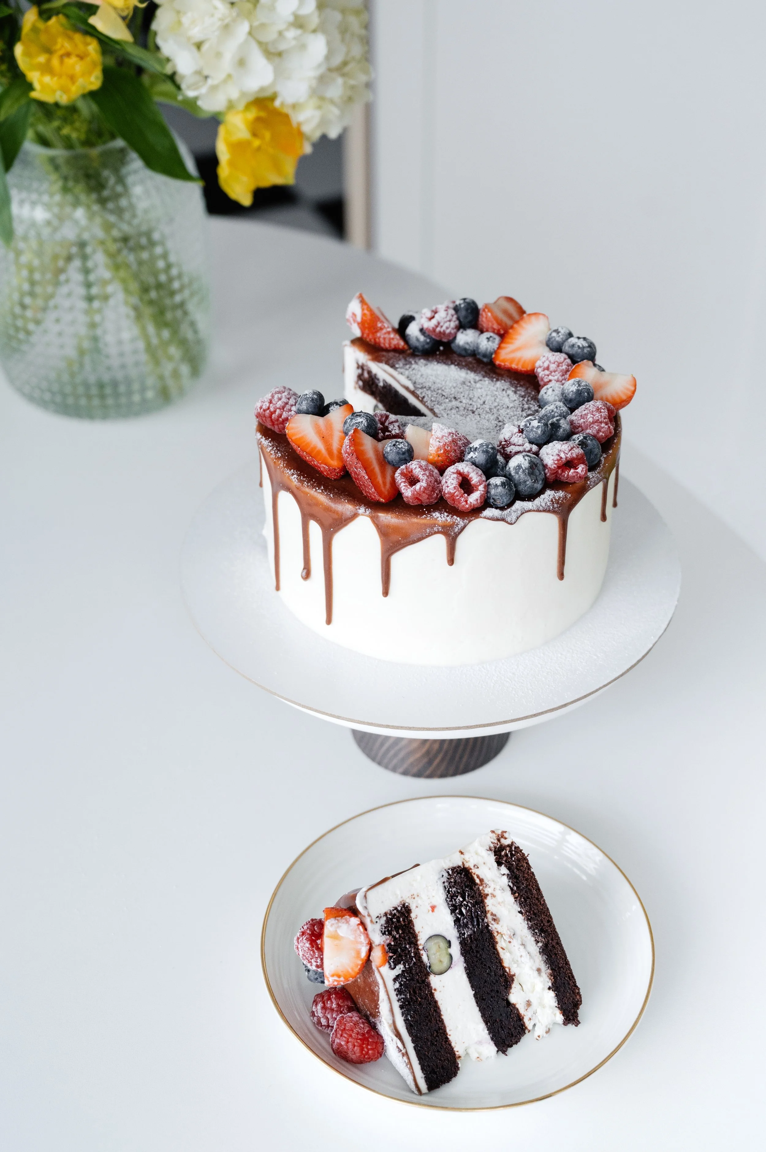 A chocolate drip cake topped with fresh strawberries, blueberries, and raspberries, with a slice removed showing layers of chocolate cake and white frosting, on a white cake stand.