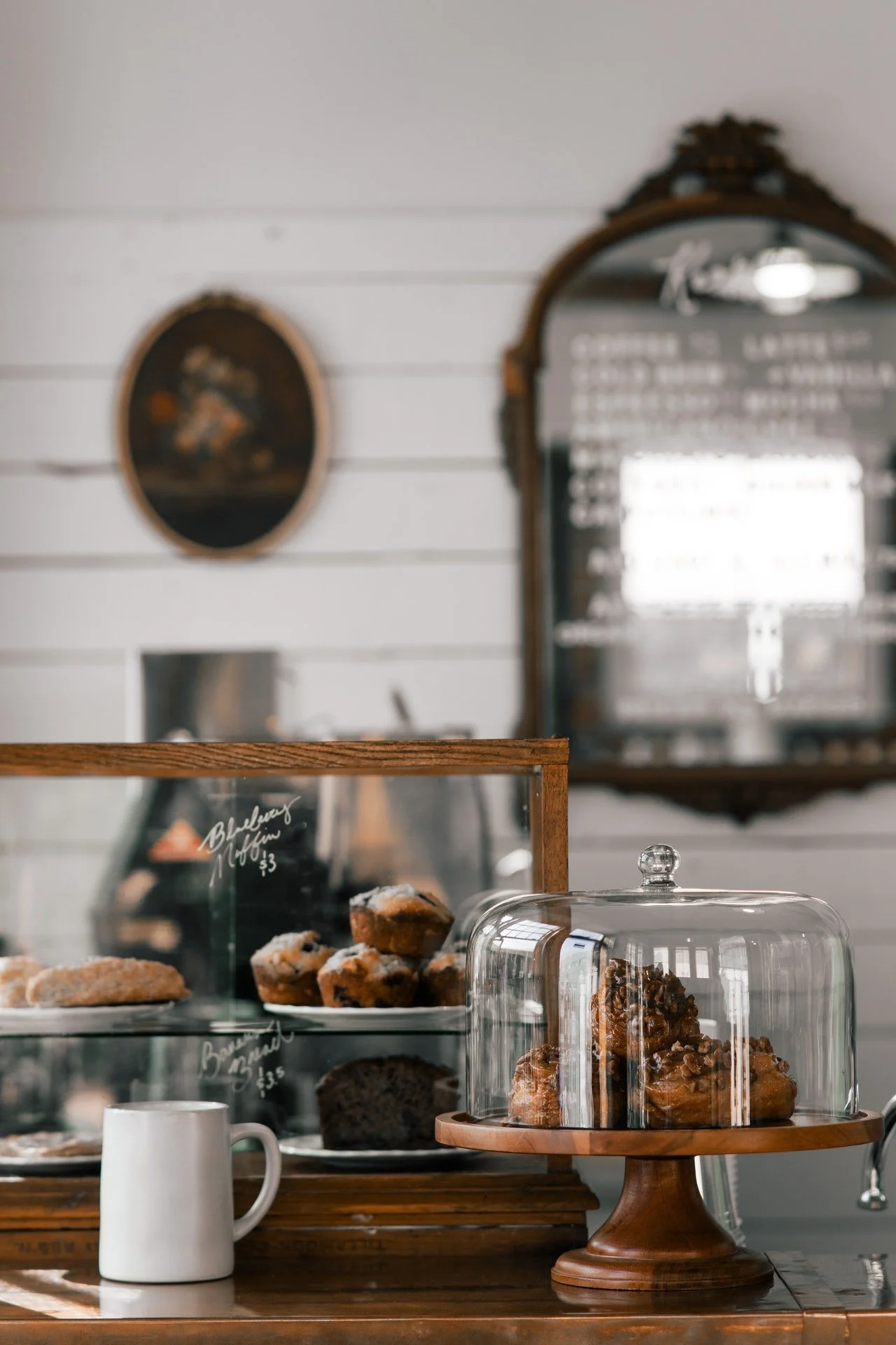 Display case with blueberry muffins and other baked goods, a glass cake dome with cookies on a wooden stand, white ceramic mug, mirror with menu and framed artwork on a white paneled wall.