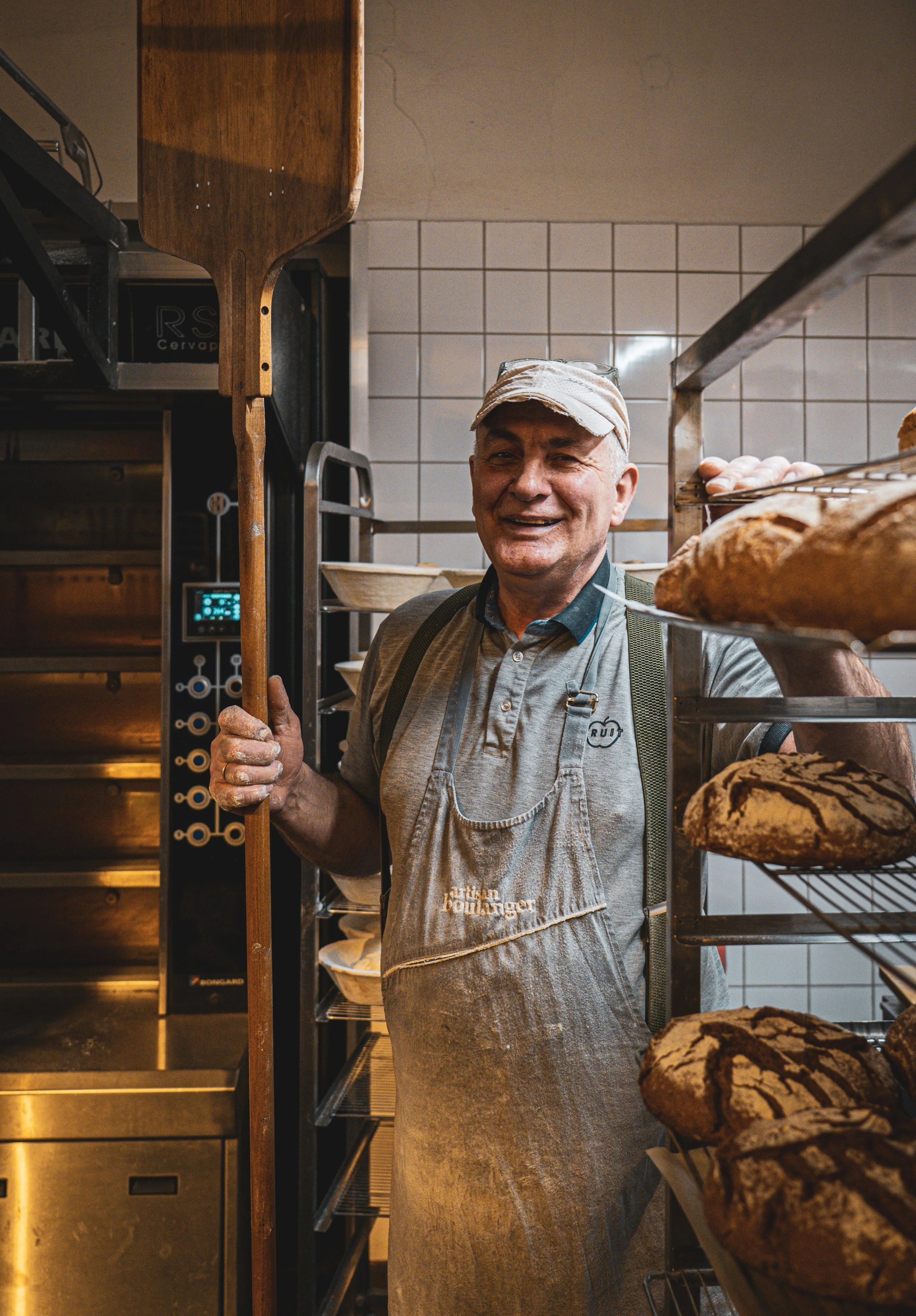 A smiling man in a baker's apron and cap standing in a bakery with racks of bread.