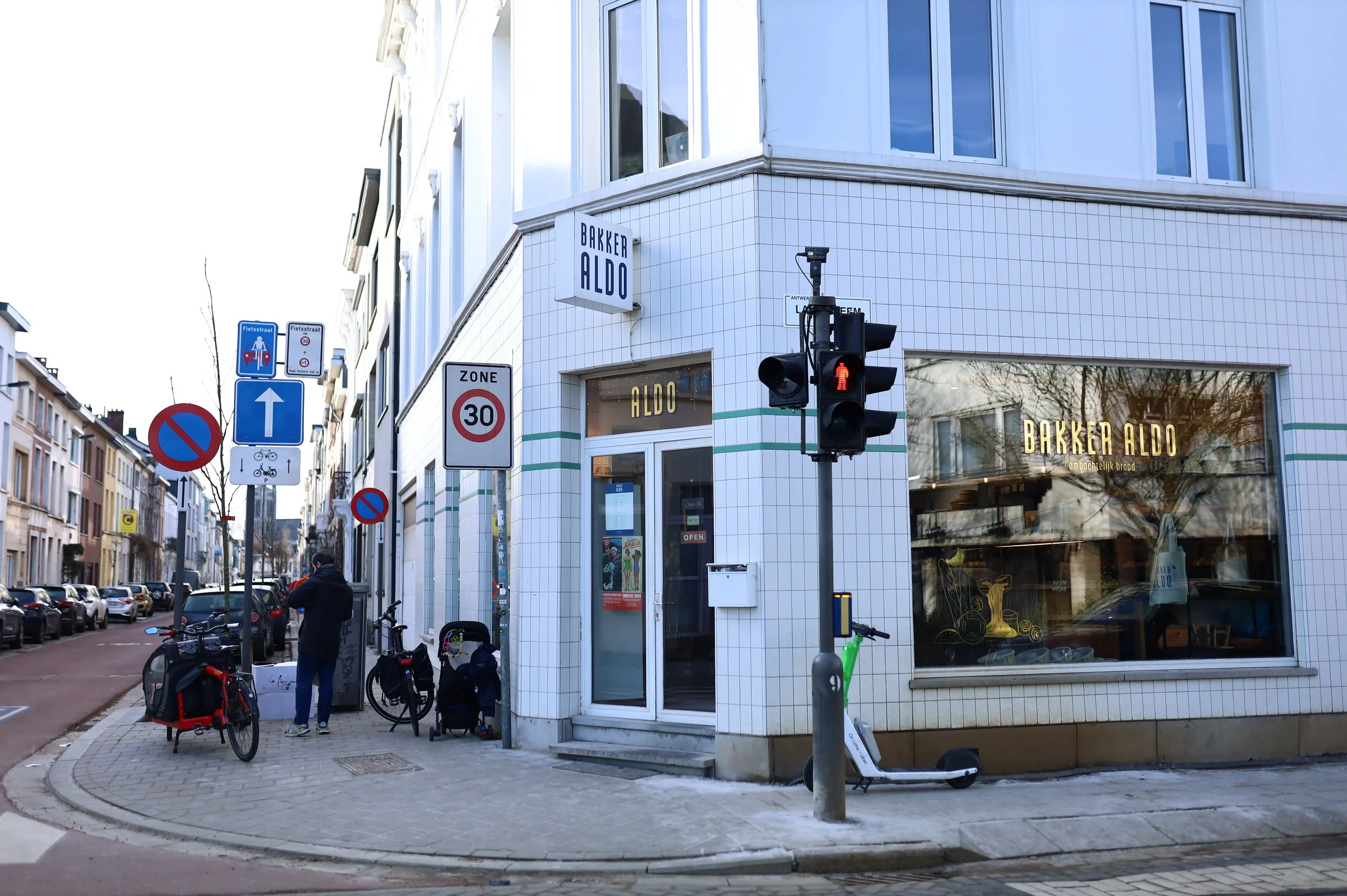 Street corner with a bakery named Bakker Aldo, bicycle, scooter, traffic lights showing red pedestrian signal, street signs indicating no parking, 30 km/h zone, and a person walking with a bicycle.