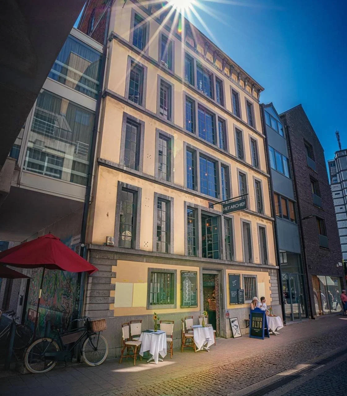 A street scene with outdoor tables and chairs in front of a multi-story building with large windows, a sign that reads 'Het Archief', and a bicycle parked on the sidewalk under a red umbrella. The building has an old architectural style with a modern adjacent building on the right, and sunlight creating lens flare in the clear blue sky.