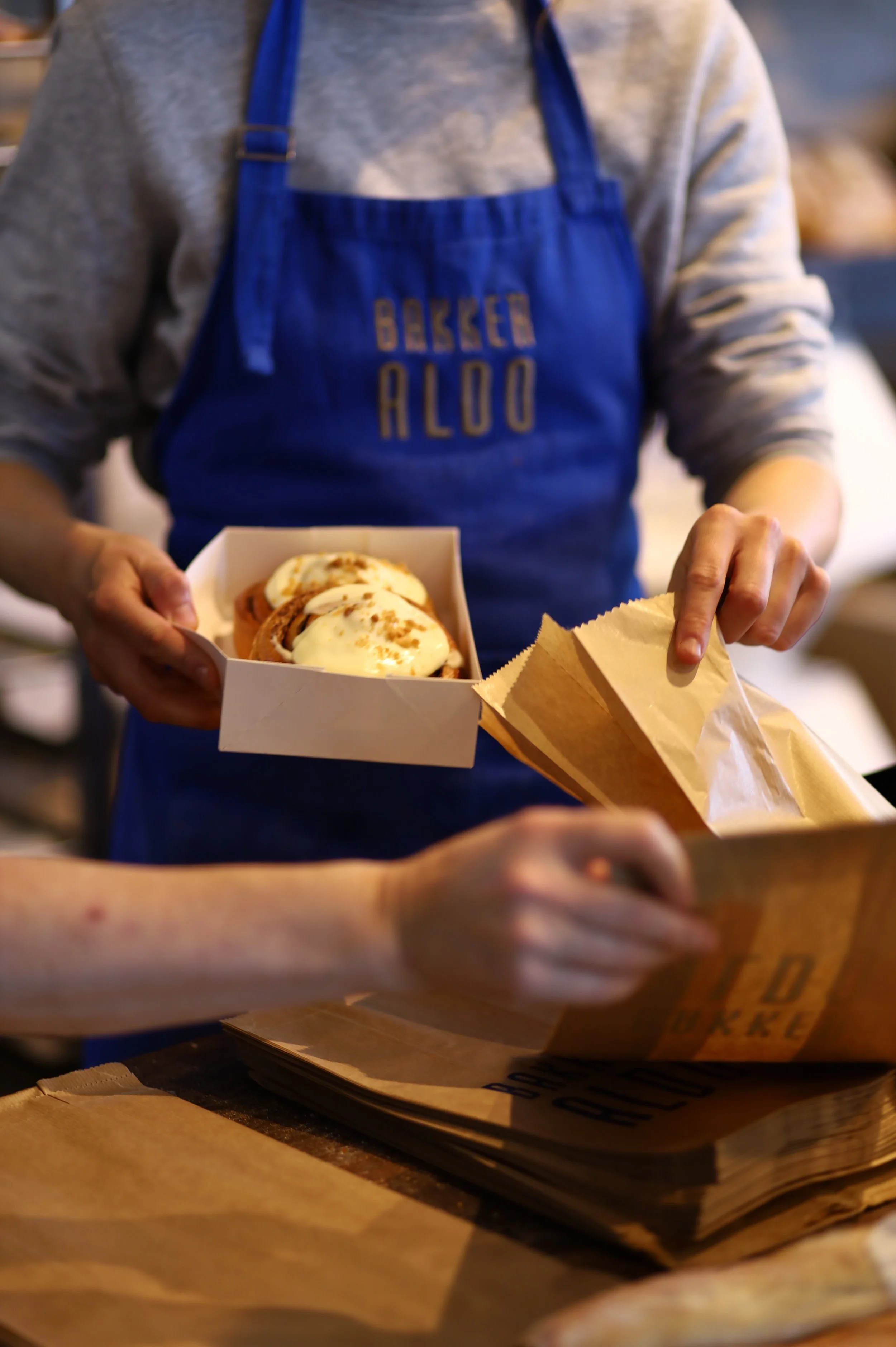 Person in a blue apron serving a box of cinnamon rolls with icing, at a bakery or café counter.