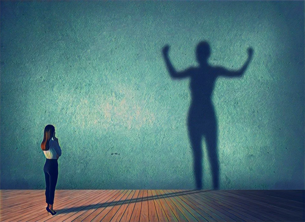 A woman gazes thoughtfully at a wall where her shadow shows her flexing confidently, symbolizing self-belief and the power of recognizing one’s value.