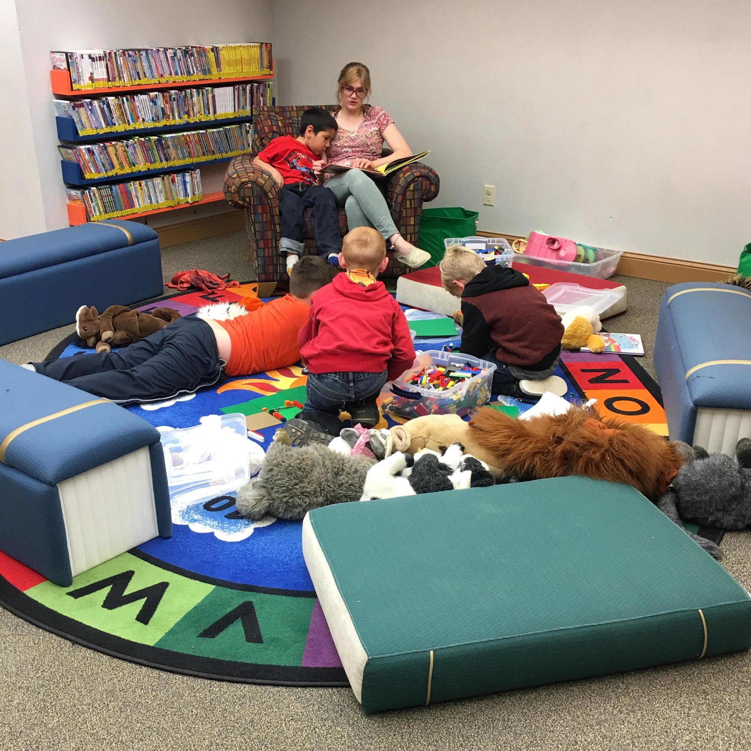 Adult reads to child as other children look on among LEGO blocks and stuffed toys.