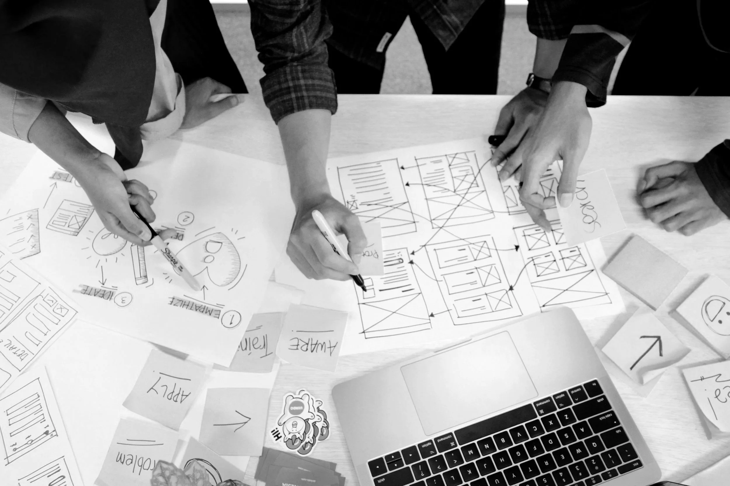 People working on a project at a table with sketches, sticky notes, a laptop, and markers.