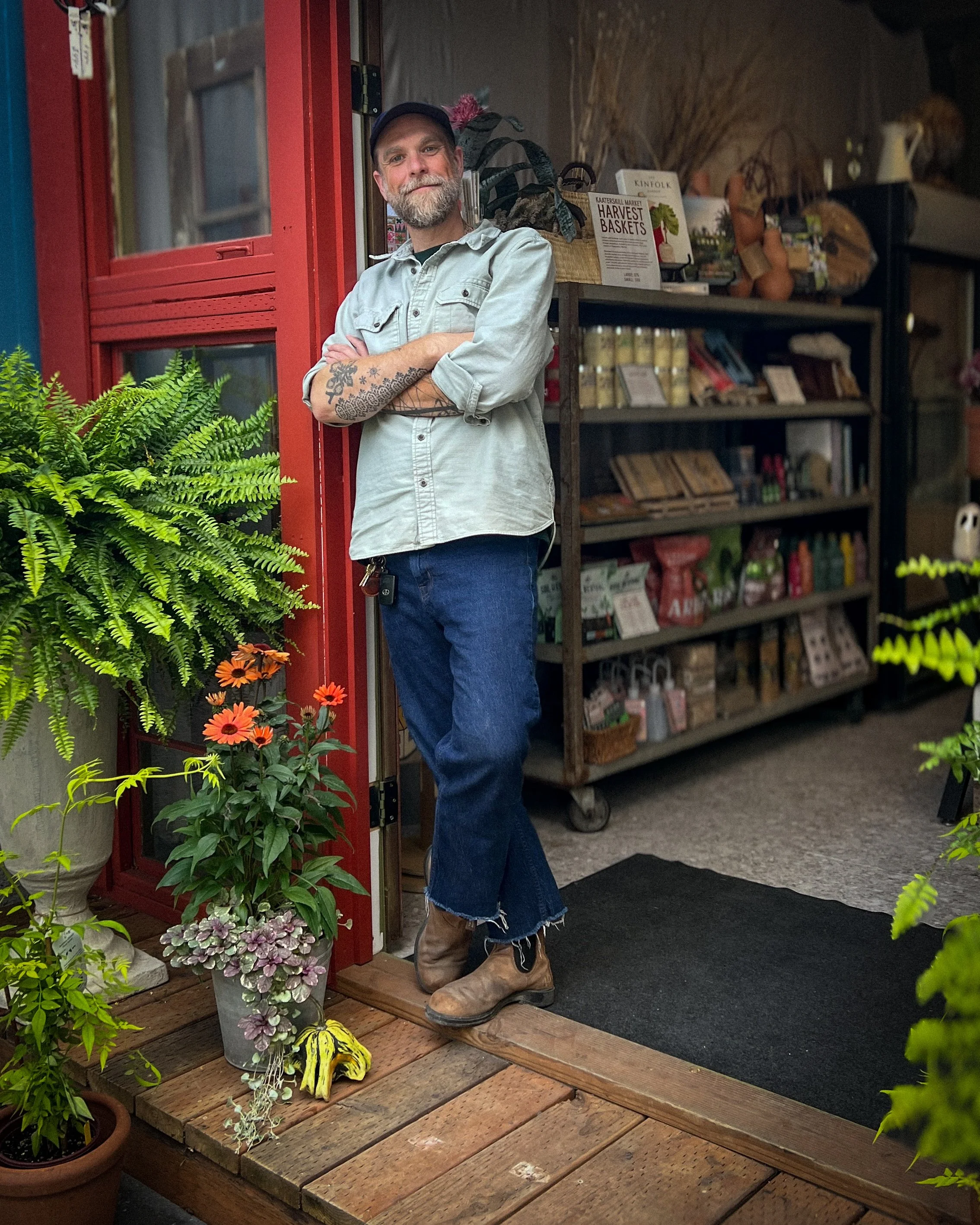Bearded man in a cap and boots standing with crossed arms at the entrance of a plant-filled market.