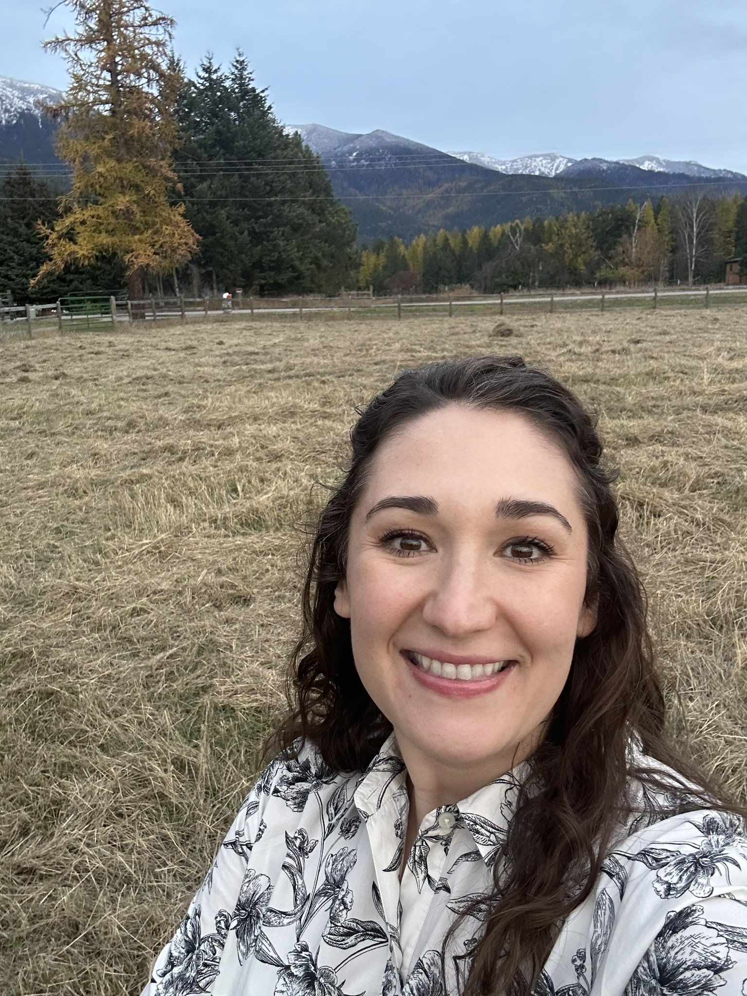 A woman with long dark hair takes a selfie outdoors in a grassy field with mountains, trees, and a fence in the background.