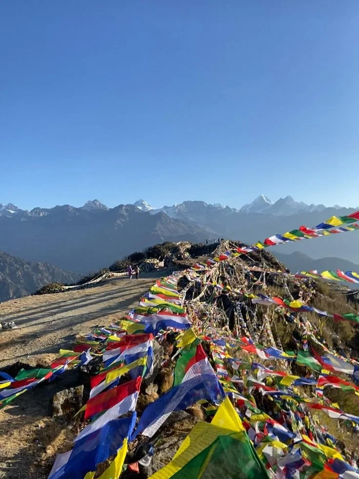 A mountain trail decorated with colorful prayer flags, with snow-capped mountains in the background and clear blue sky overhead.