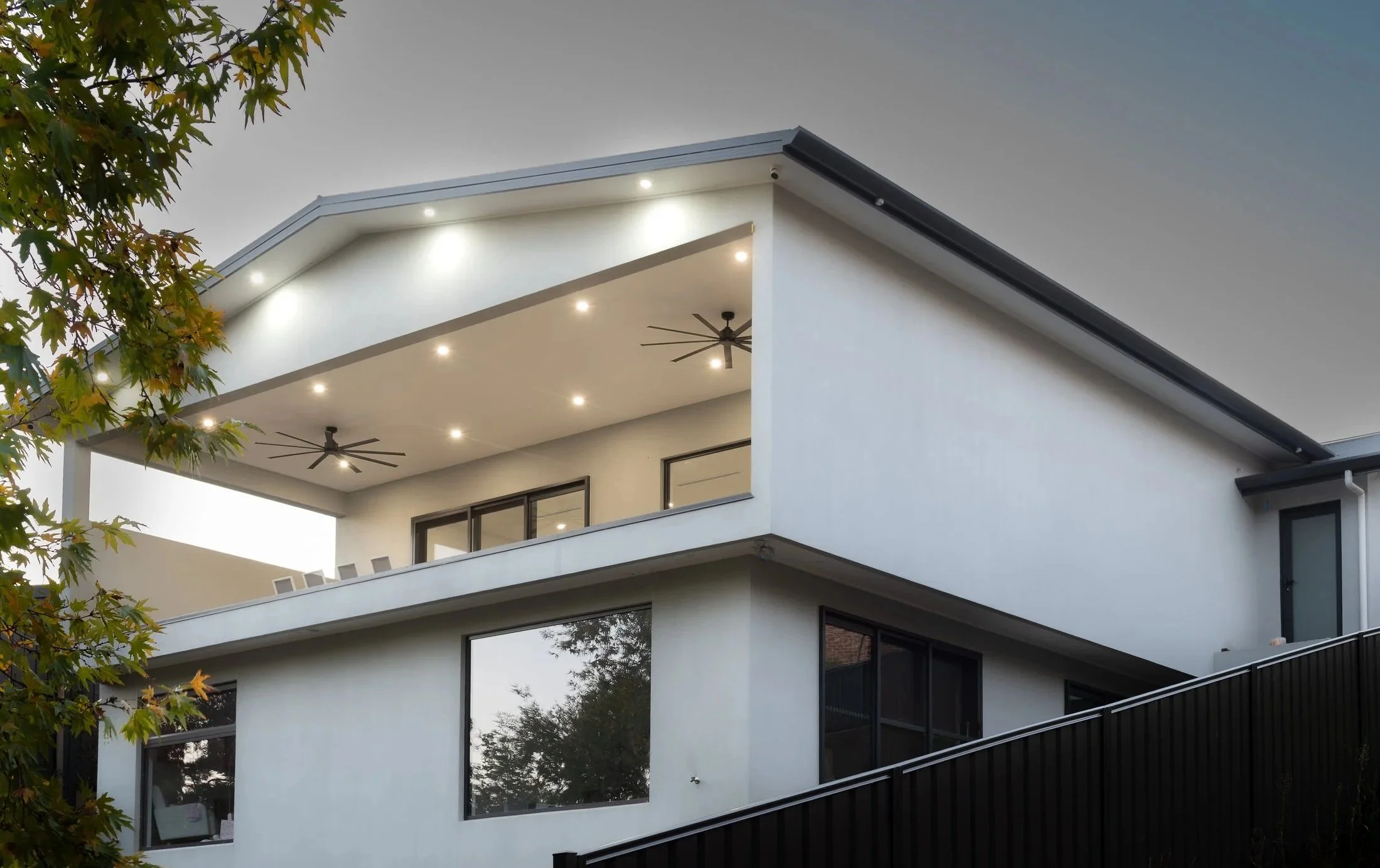 Modern two-story house with a large balcony on the upper floor, white exterior walls, large windows, and a partial black fence on a slightly sloped yard with a tree in the foreground.