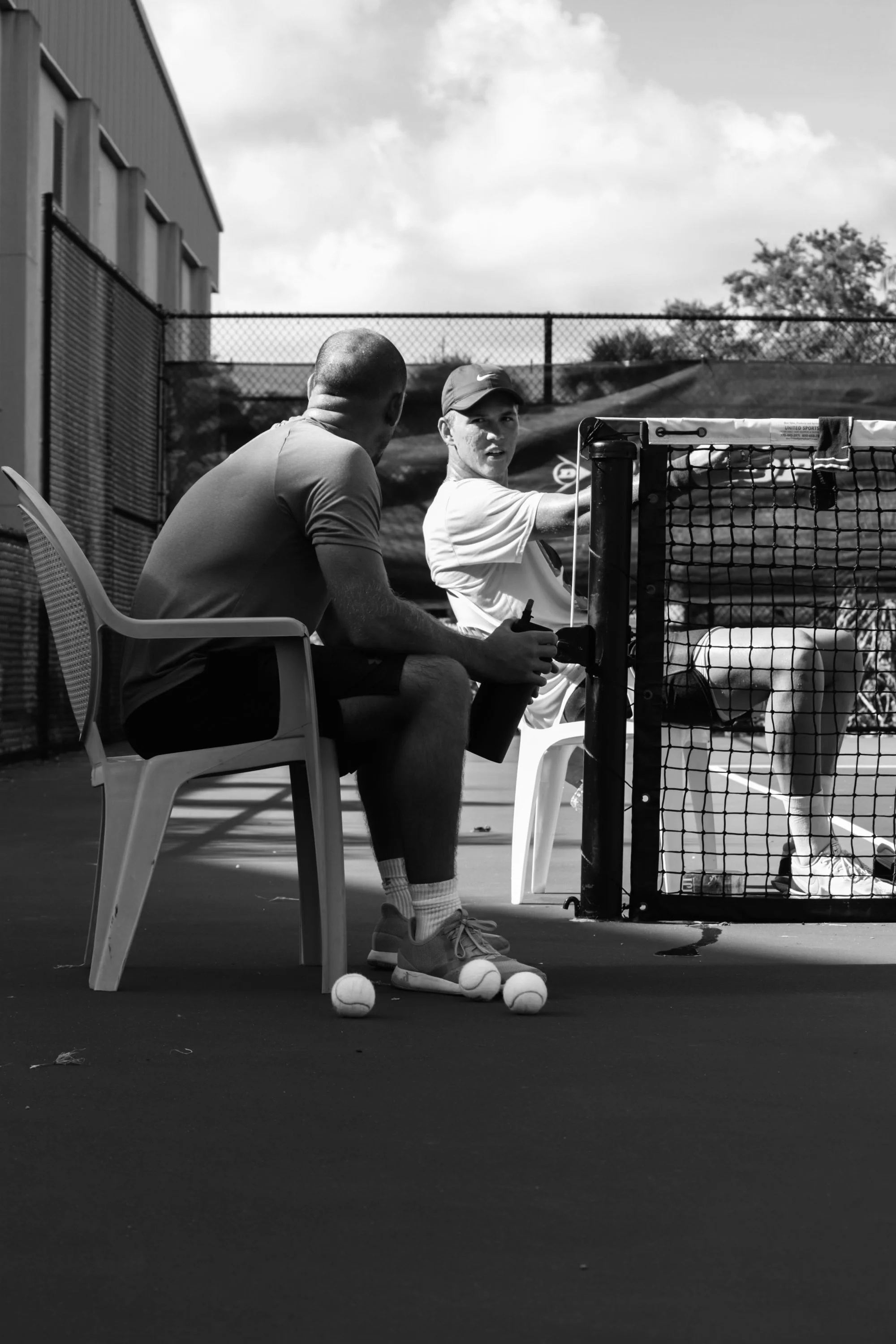 Two men sitting by a tennis court, engaged in conversation; one is wearing a cap and sportswear, the other has a closely shaved head and T-shirt; tennis balls and racquets are visible.