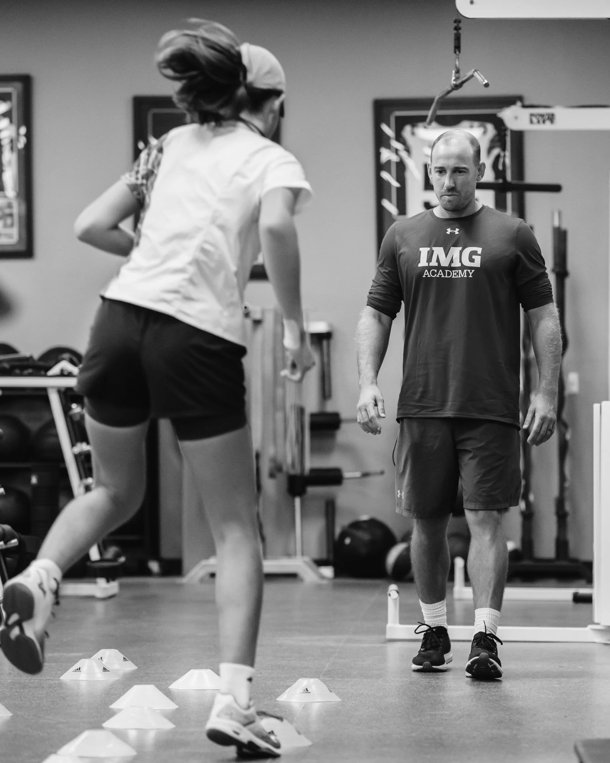 A man and a woman working out in a gym. The woman is running around orange cones, while the man watches. The man is wearing an 'IMG Academy' shirt, shorts, and sneakers. The gym has fitness equipment and framed pictures on the walls.