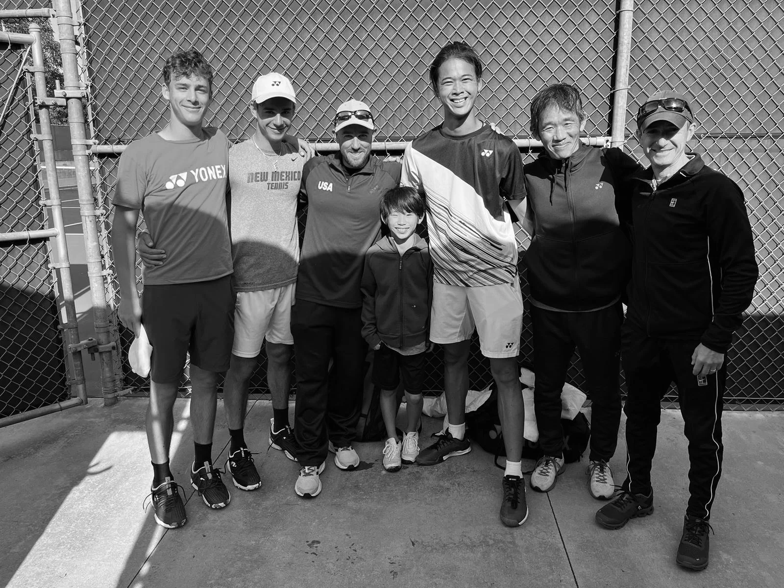 Group of eight people, including young and adult individuals, standing together on a tennis court in front of a chain-link fence. They are dressed in athletic clothing, some with tennis gear, and are smiling.