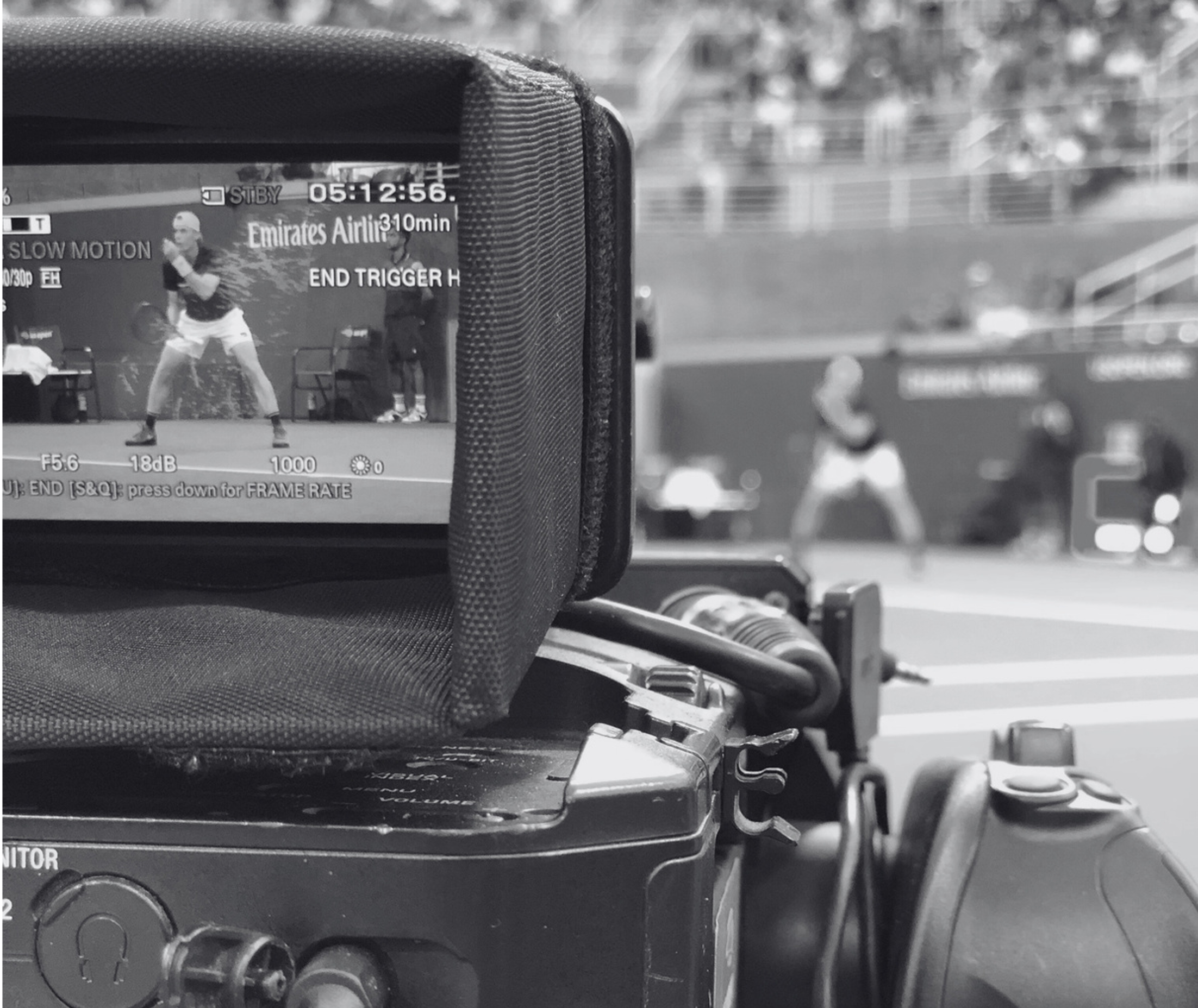 Close-up of a camera screen showing a tennis player in action, with part of the camera body and some equipment in the foreground, and blurred tennis court background.