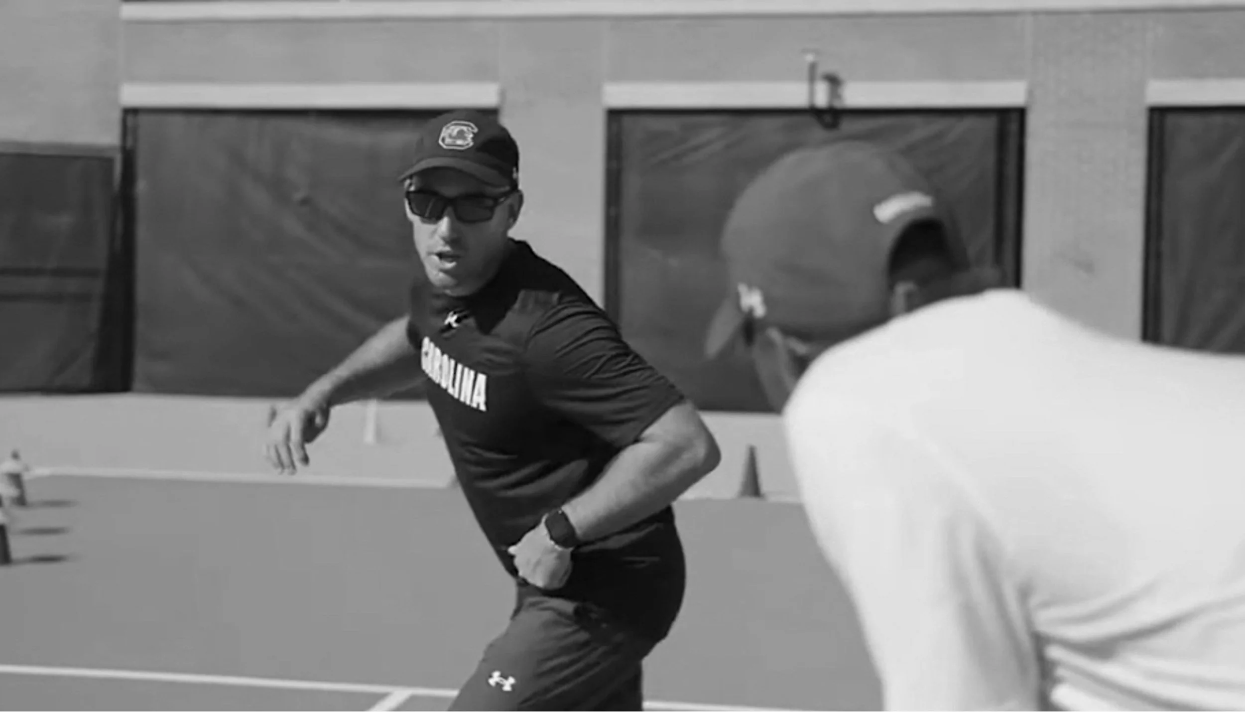 A male tennis player wearing sunglasses, a black cap, and a black shirt with the word 'Carolina' on it, is in motion on a tennis court during a match, with a spectator in a gray shirt and cap watching nearby.