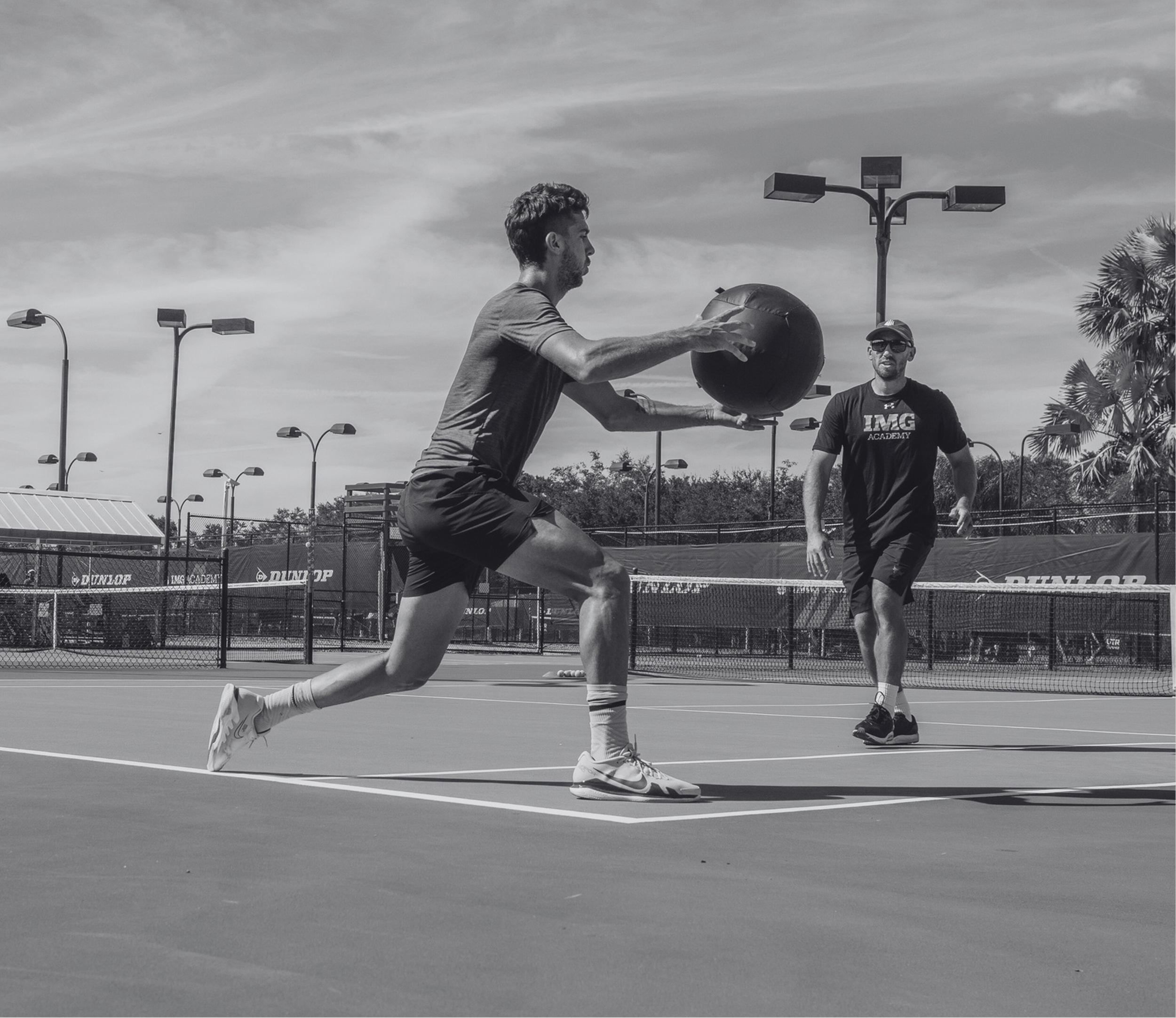 A young man playing tennis on an outdoor court, reaching to hit a ball with a racket, while another man stands nearby watching. The scene is sunny with multiple light poles, and palm trees can be seen on the side.