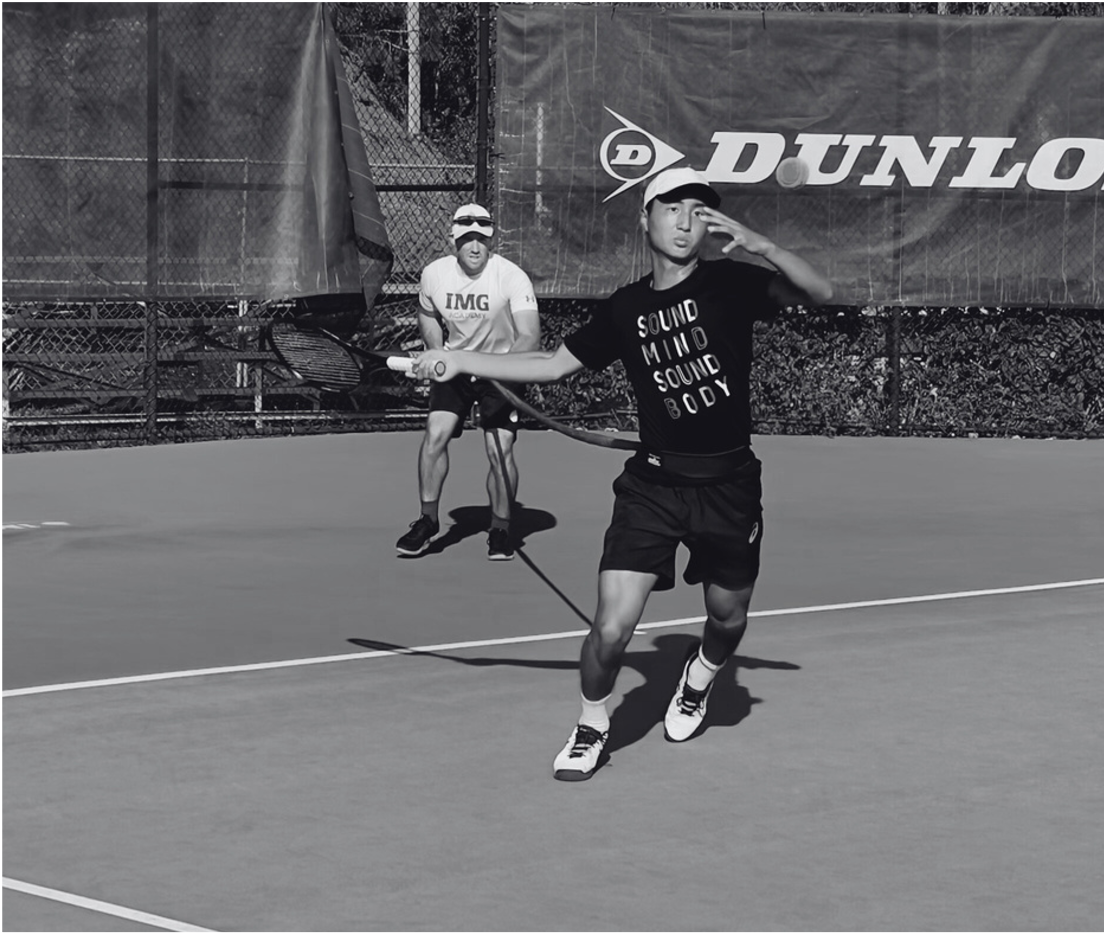 A young man playing tennis on an outdoor court, wearing a black t-shirt and shorts, with a tennis racket in his hand. Another man in the background is wearing a white shirt and cap, sitting near the fence. There is a large banner with the Dunlop logo