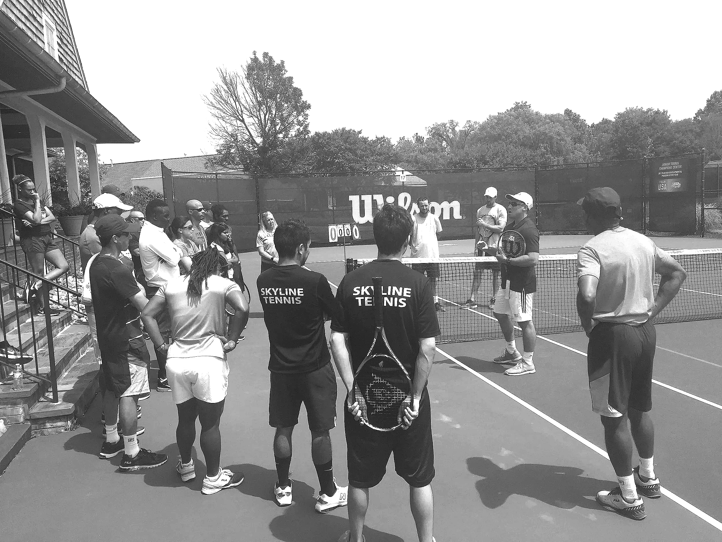 A group of people gathered on a tennis court, with some standing around and others holding tennis rackets, possibly in a tennis lesson or training session.