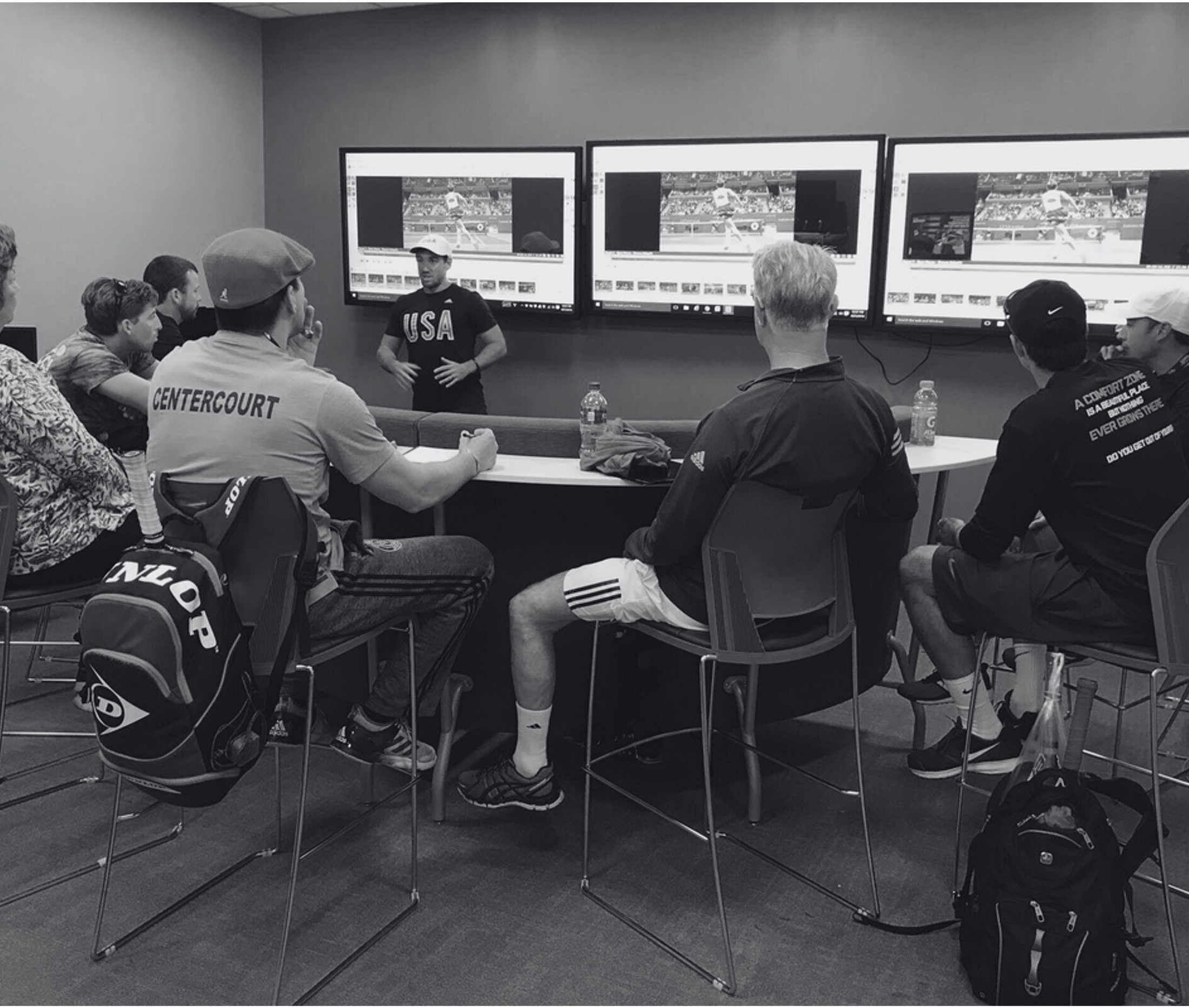 A group of people sitting and listening to a man giving a presentation in front of three large screens displaying tennis match footage. The attendees are casually dressed and are seated at tables with water bottles.