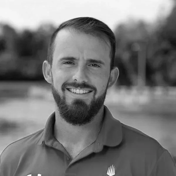 Black and white photo of a smiling man with a beard and short hair, wearing a polo shirt, outdoors.