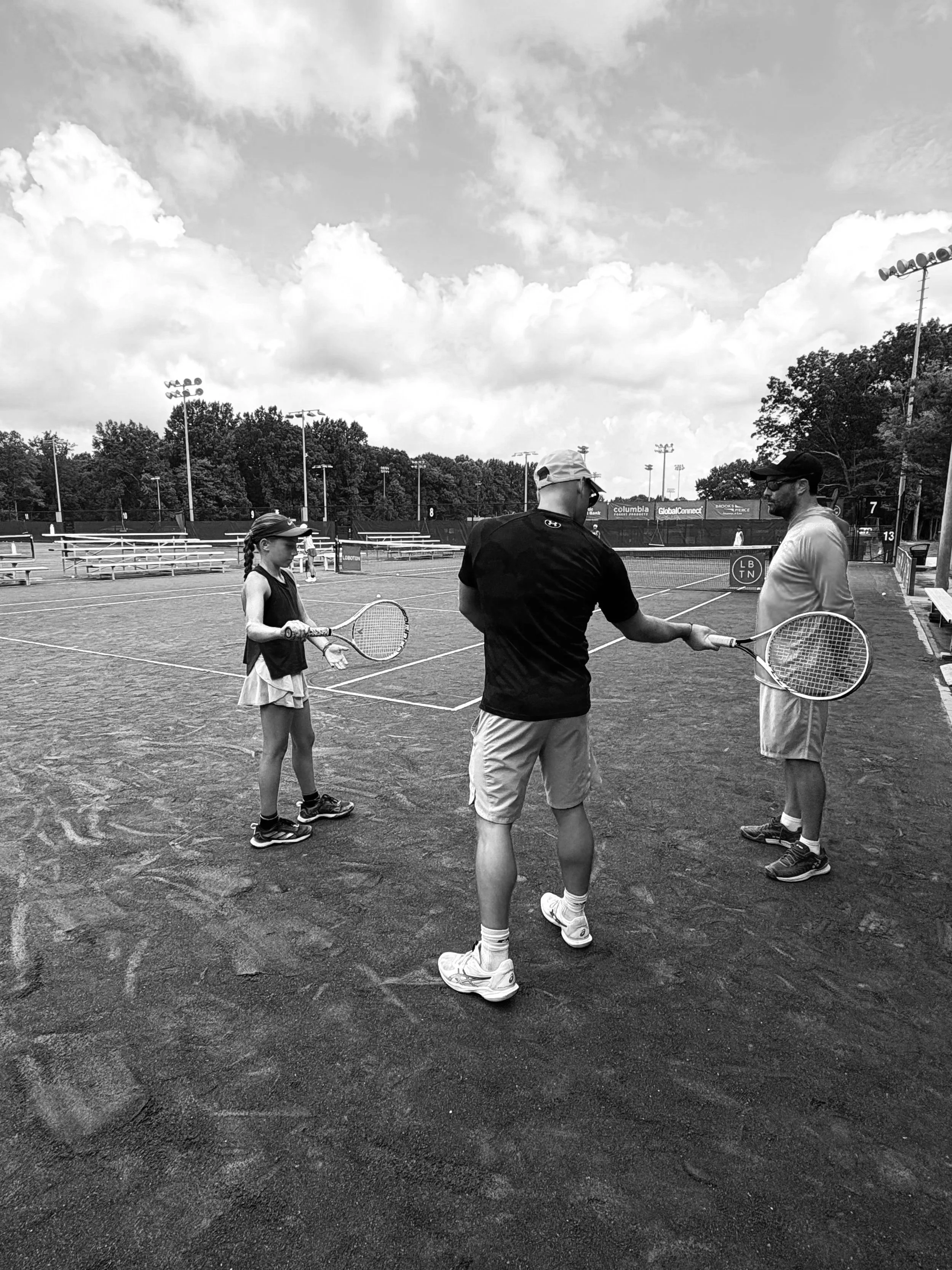 Two children and two adults on a tennis court, holding tennis rackets, with a background of cloudy sky and trees.