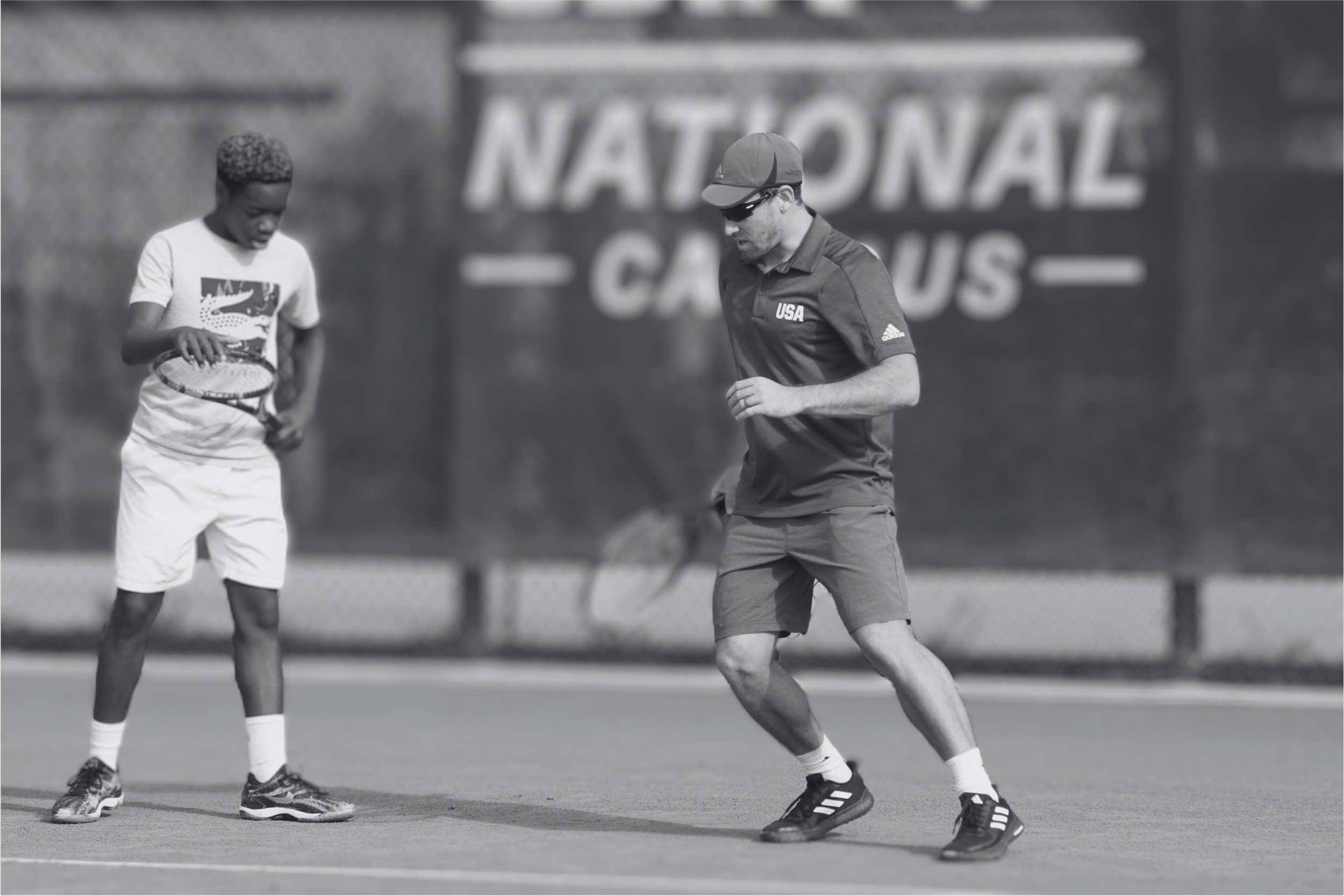 Two tennis players, one young male and one adult male, are on a tennis court, preparing to play. The adult male, wearing sunglasses, a cap, and shorts with a 'USA' logo, is holding a tennis racket while the young male is looking down at his racket. I