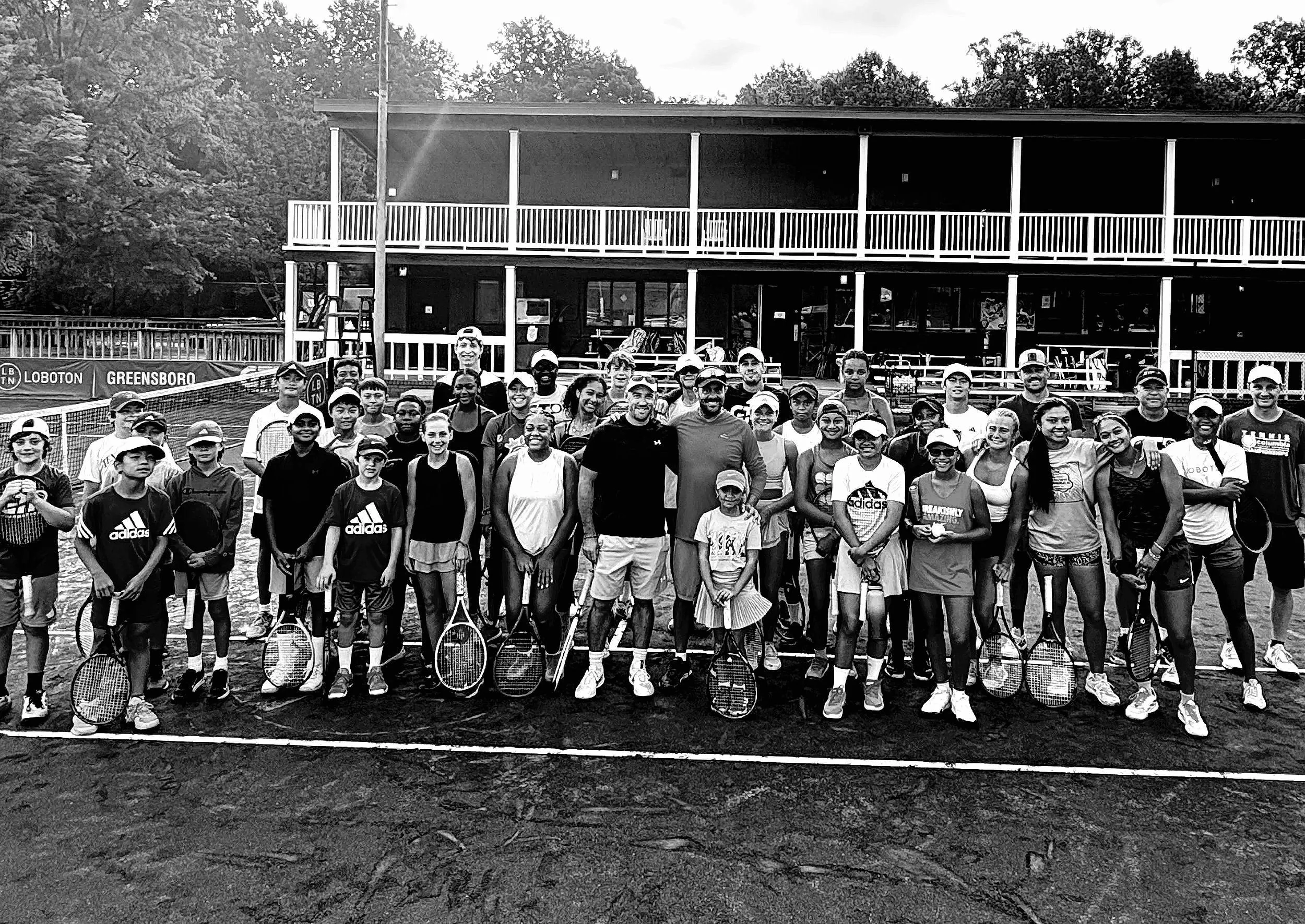 A group of people, including children and adults, standing on a tennis court with rackets, smiling at the camera, with a building and trees in the background.