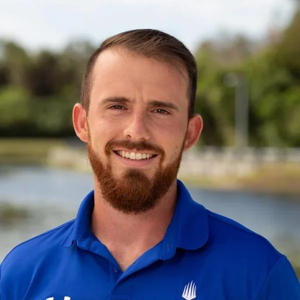 Young man with a beard smiling outdoors, wearing a blue polo shirt with a logo, in front of a blurred background with trees and a body of water.