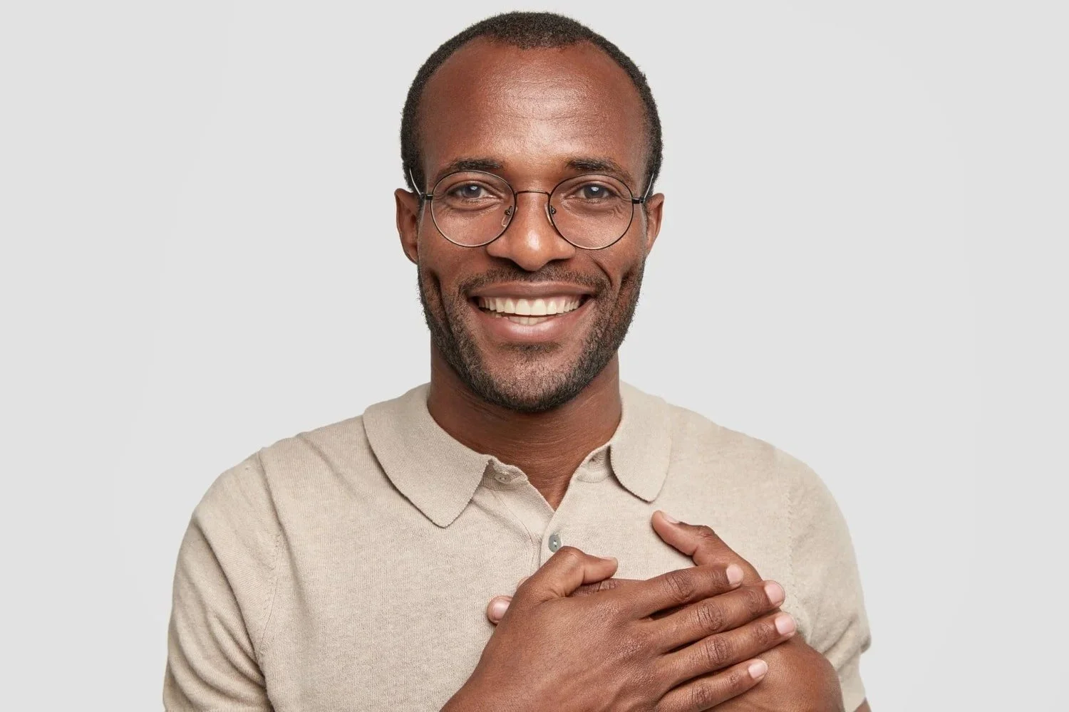 Smiling man with glasses in a beige polo shirt, holding his chest with one hand, against a light gray background.