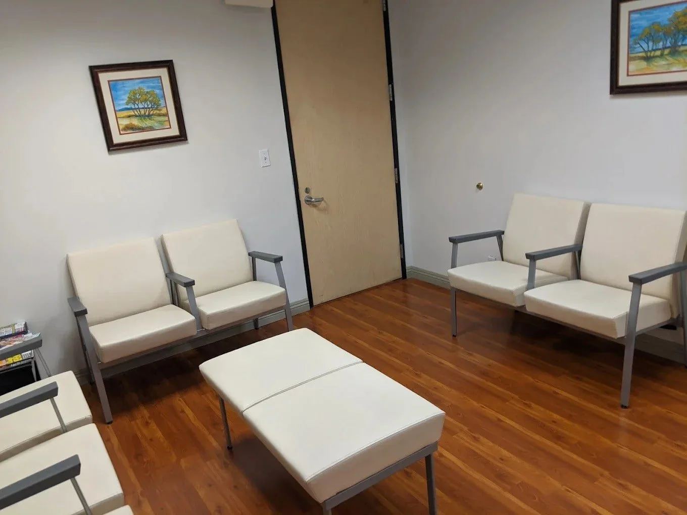 A waiting room with beige chairs with gray armrests arranged against white walls, a wooden floor, a closed wooden door, framed landscape paintings of trees and fields on the walls, and a small table with magazines.