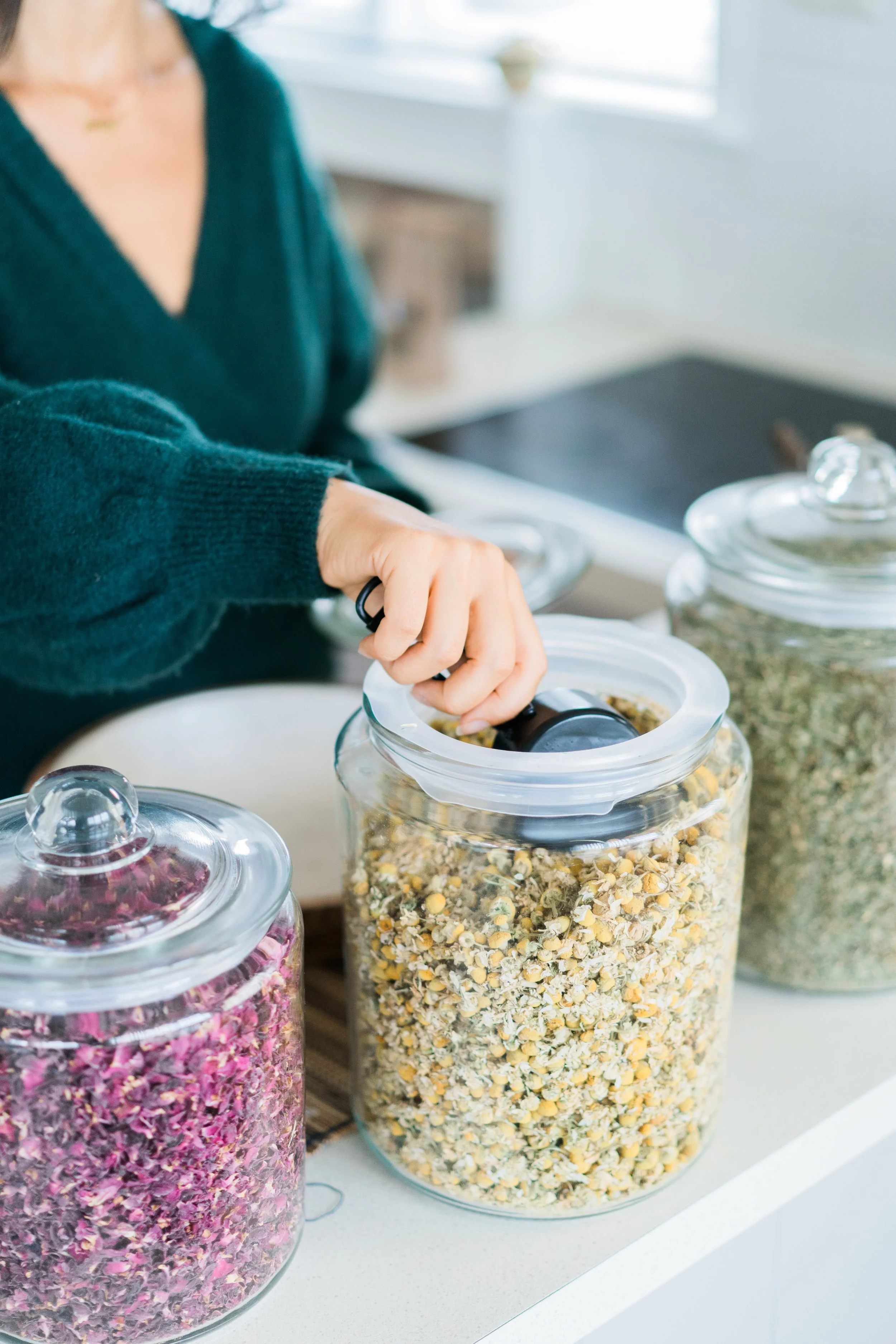 Person opening a large glass jar filled with dried chamomile flowers in a kitchen.