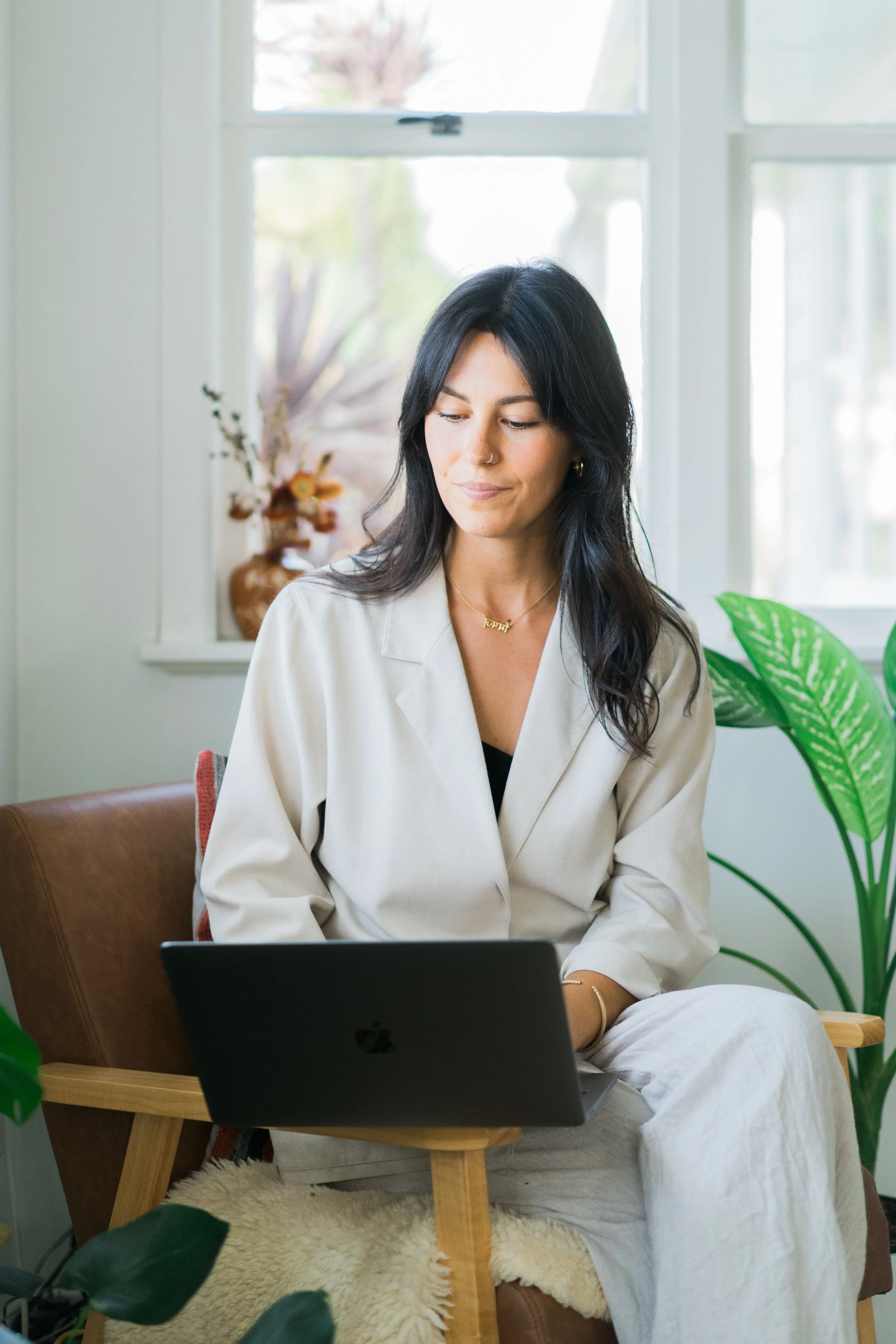 A woman with long dark hair sits in an armchair working on a laptop in a bright room with plants and a window in the background.