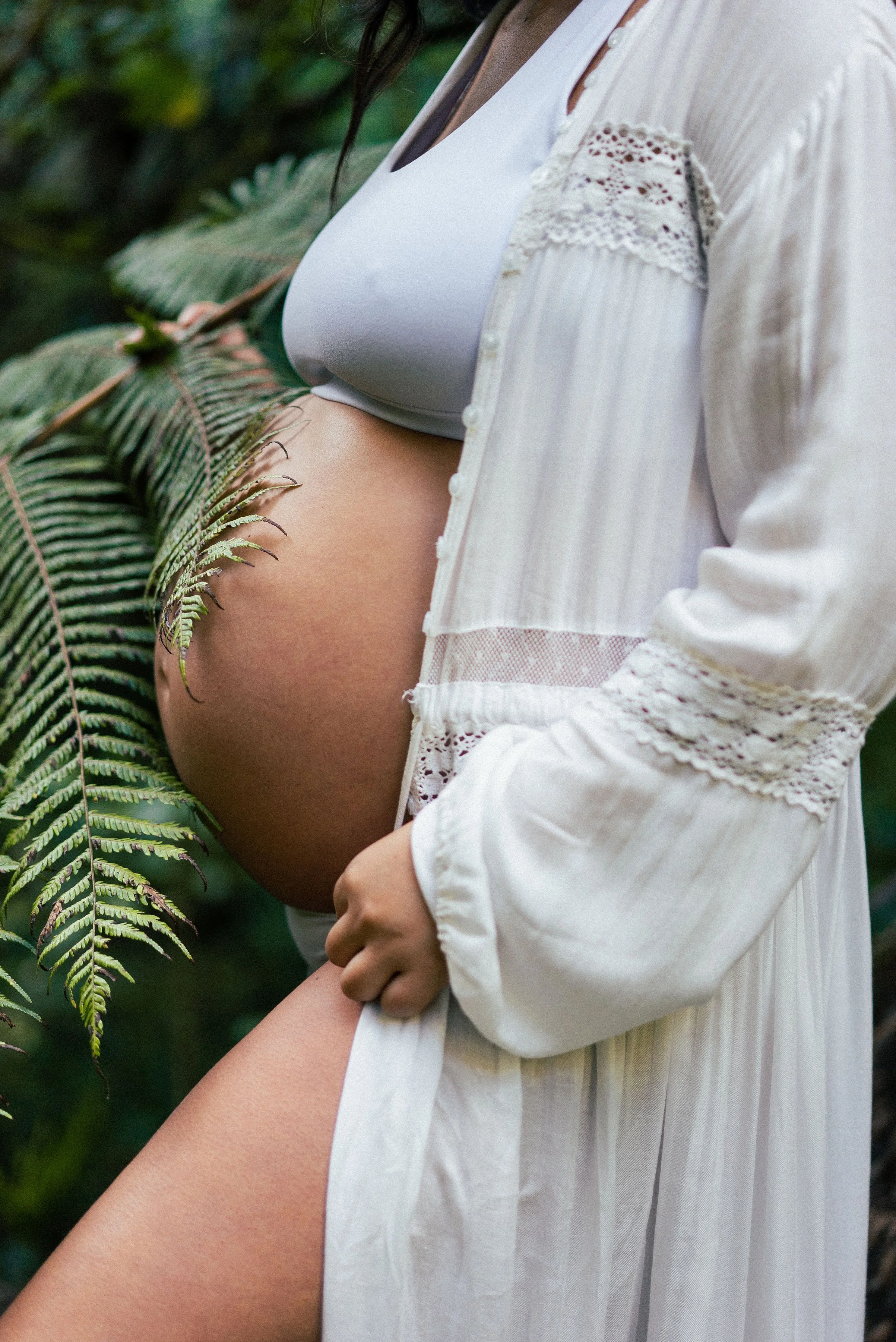 A pregnant woman gently holding her belly outdoors, surrounded by lush green foliage.