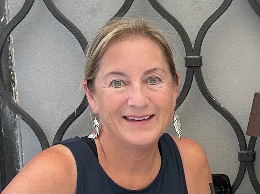 A smiling woman with short blonde hair, wearing a sleeveless navy blue top and feather-shaped earrings, sitting in front of a decorative dark metal wall.