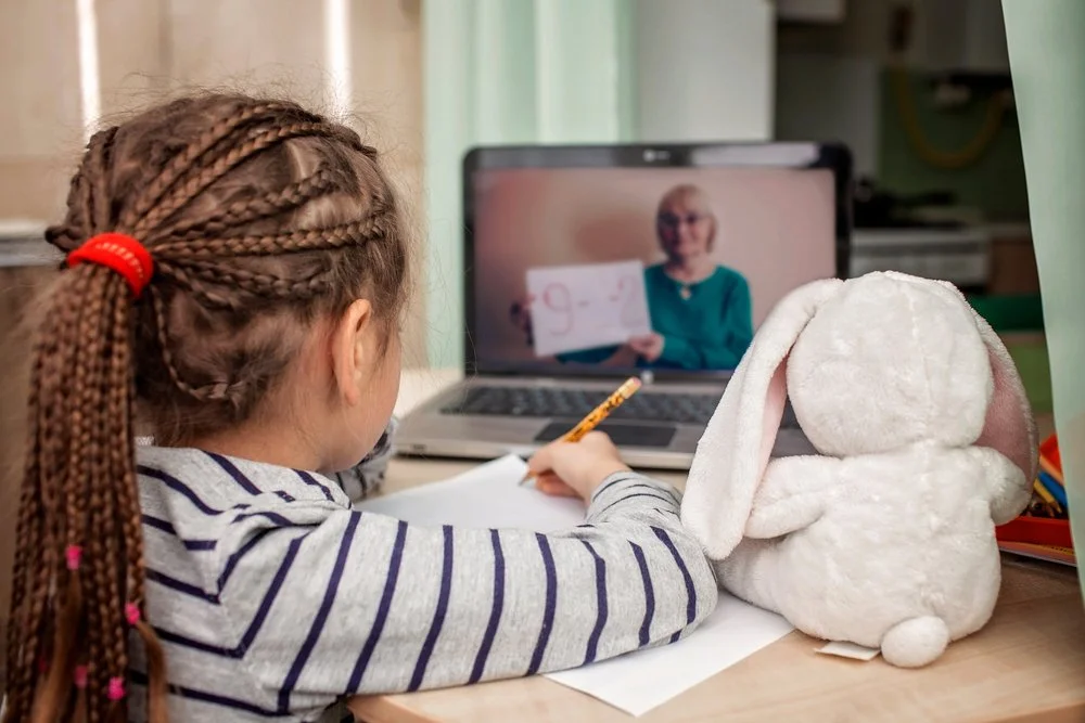 A young girl with braided hair, wearing a striped shirt, is doing homework while participating in a video call with an older woman on her laptop. A plush bunny with long ears is sitting beside her.