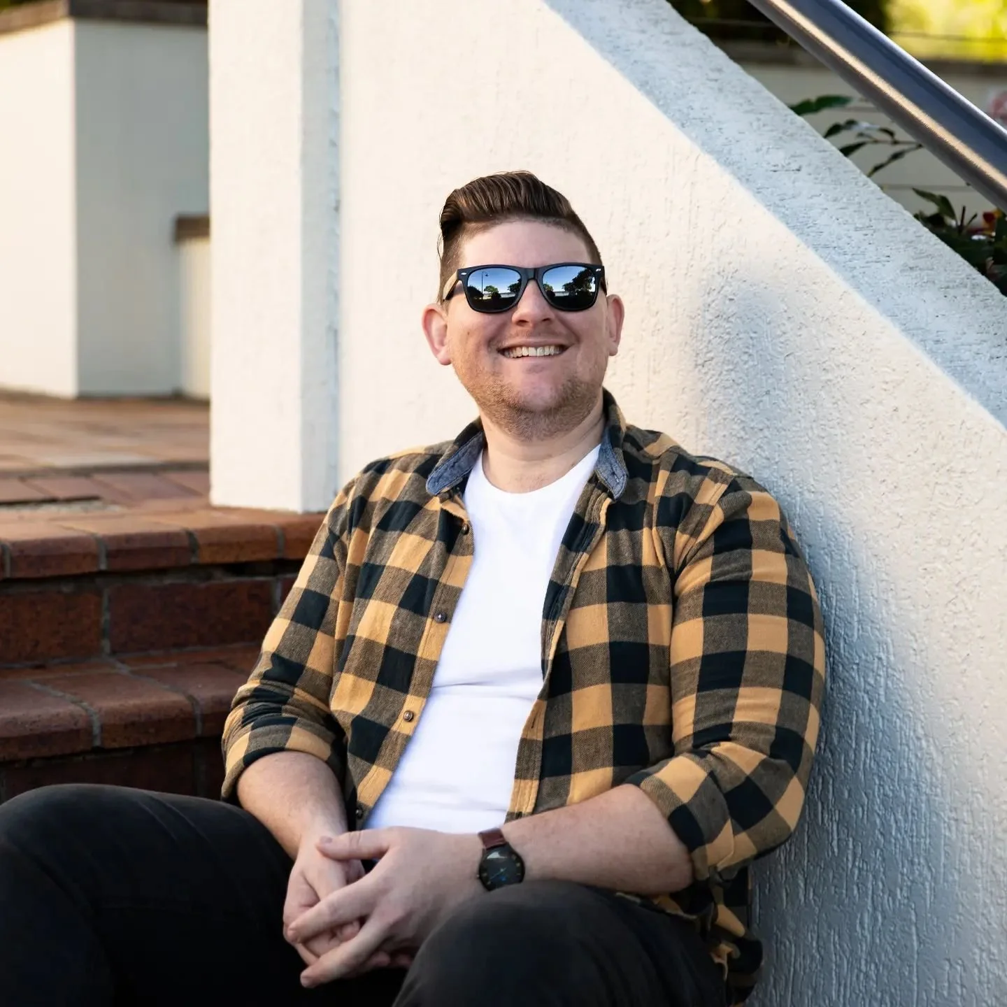 A man with short brown hair, wearing black sunglasses, a yellow and black plaid shirt over a white t-shirt, smiling, sitting outside against a white textured wall with brick stairs behind him.