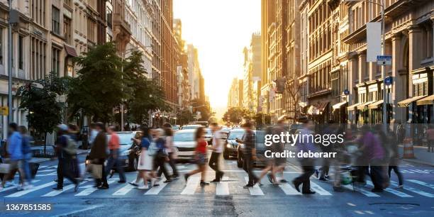 A busy city street at sunset with pedestrians crossing and cars driving by, surrounded by tall buildings.