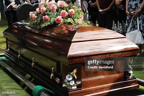 A wooden casket with a floral arrangement on top, surrounded by people at a funeral service.