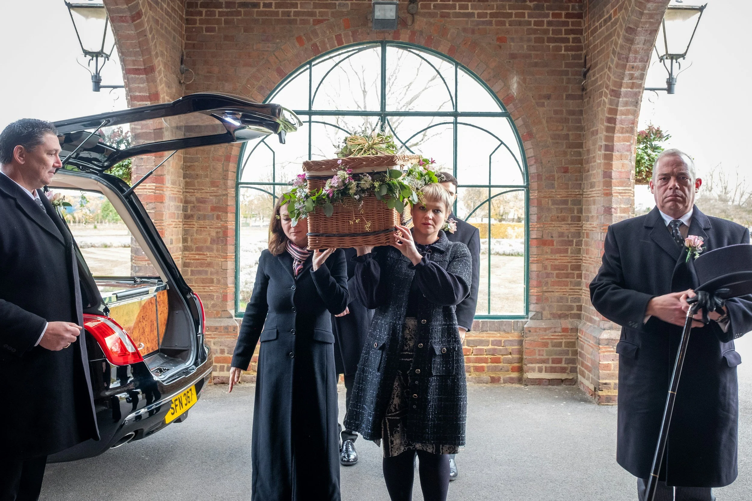 A woman carrying a basket of flowers during a funeral procession, with a hearse and pallbearers in formal attire in the background inside a brick building with large arched windows.