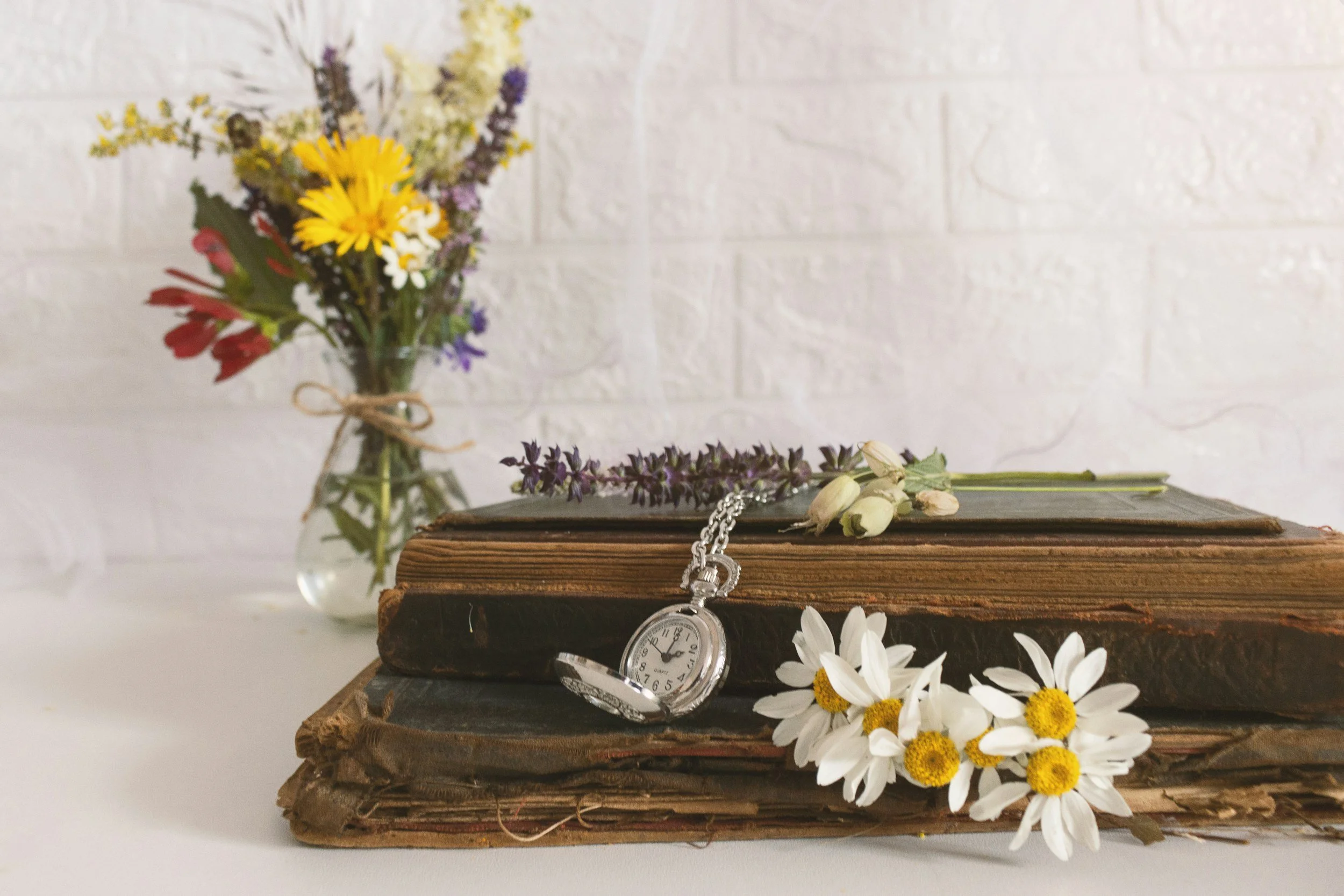 Still life of two old books with worn pages, a pocket watch, and small white daisies with yellow centers, with a glass vase of colorful wildflowers in the background against a white brick wall.